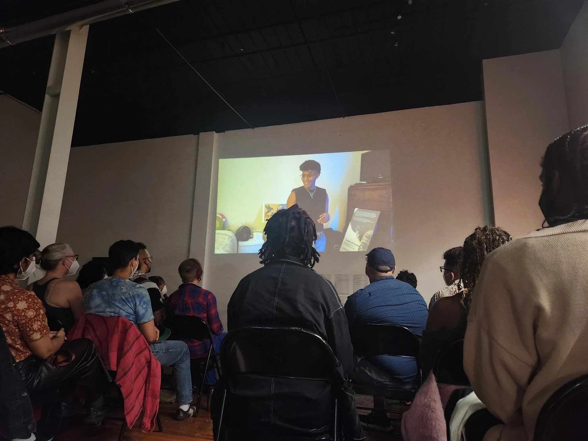 A crowd watching BE HERE NOW on a large screen, The Clown visible.