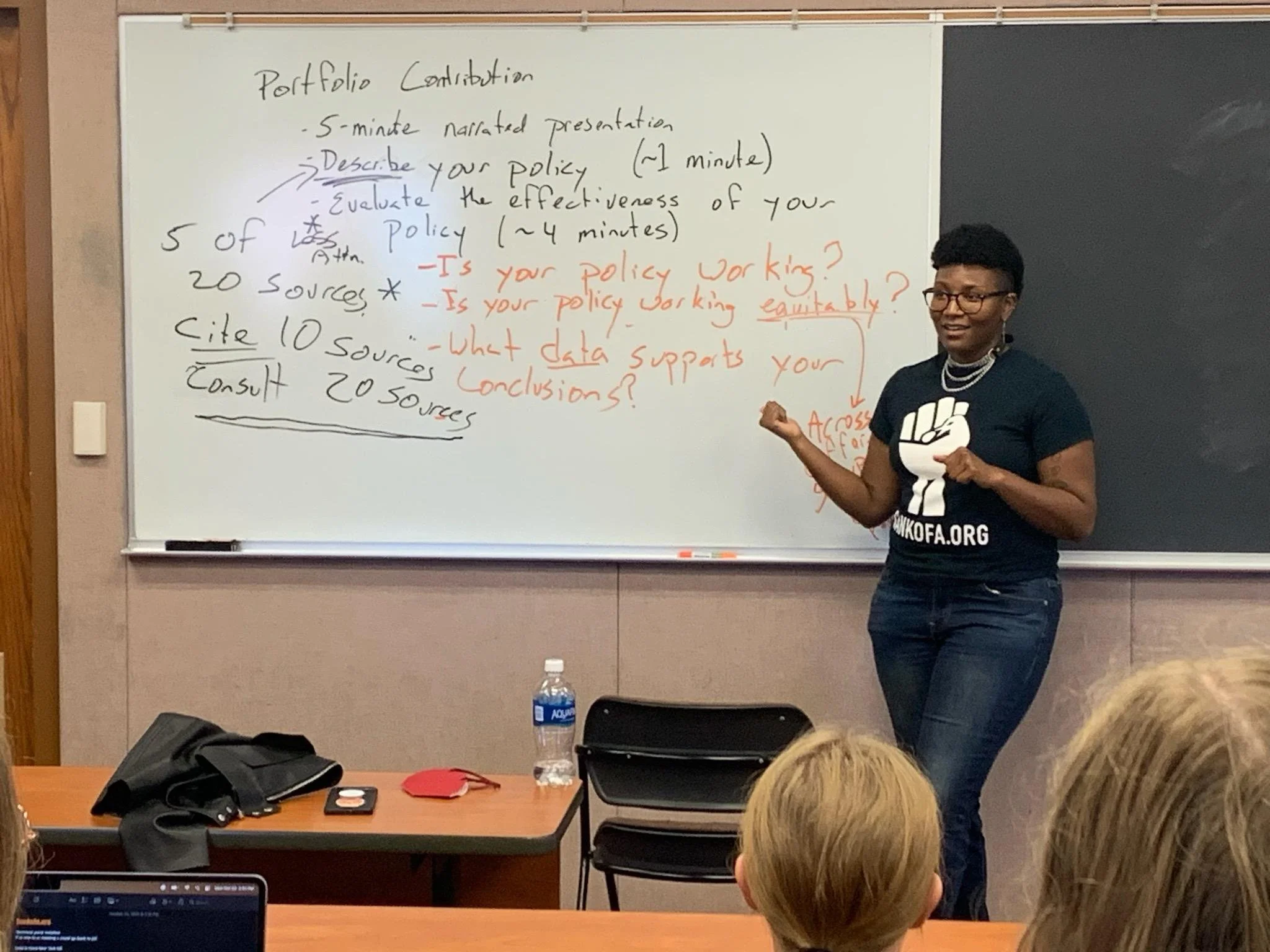 Kenyatta in front of a classroom. They are wearing a black shirt with a fist on it. Image by Dennis Plane, 2023