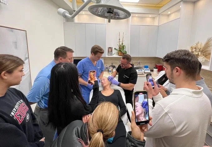 Group observing a medical procedure in a clinic setting, focused on a woman receiving facial treatment, with multiple people taking photos on smartphones.