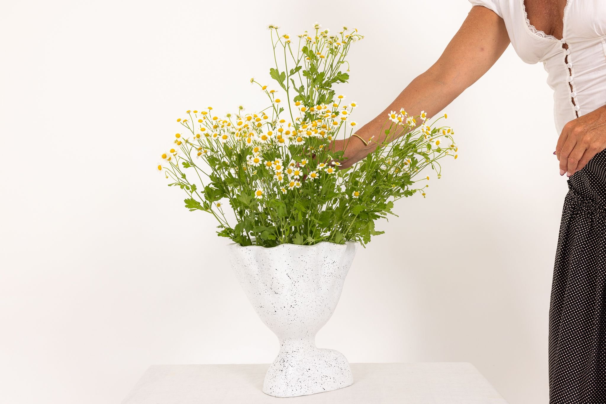 Person arranging a large white textured flower vase with yellow and white daisies on a white surface against a plain white background.