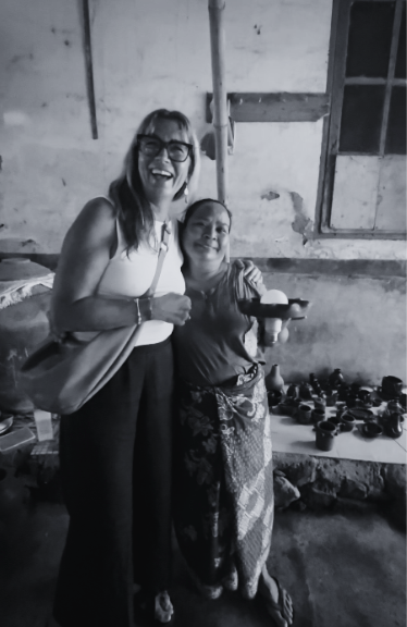 Two women smiling together indoors, one holding a small bowl. Various clay pots on a shelf in the background. Black and white photo.