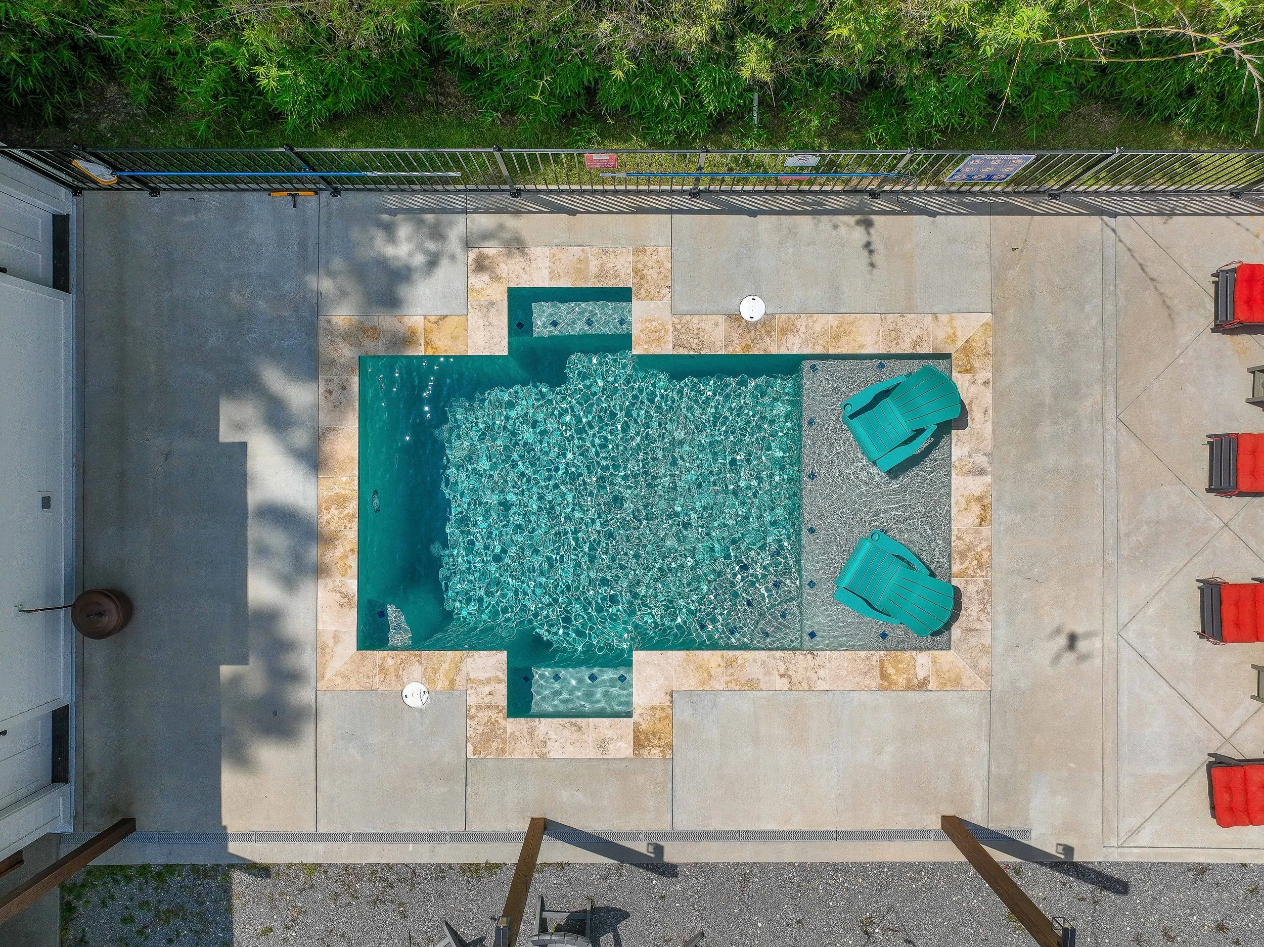 A top-down view of a backyard patio with a swimming pool, green chairs, a fence, and some trees and bushes.