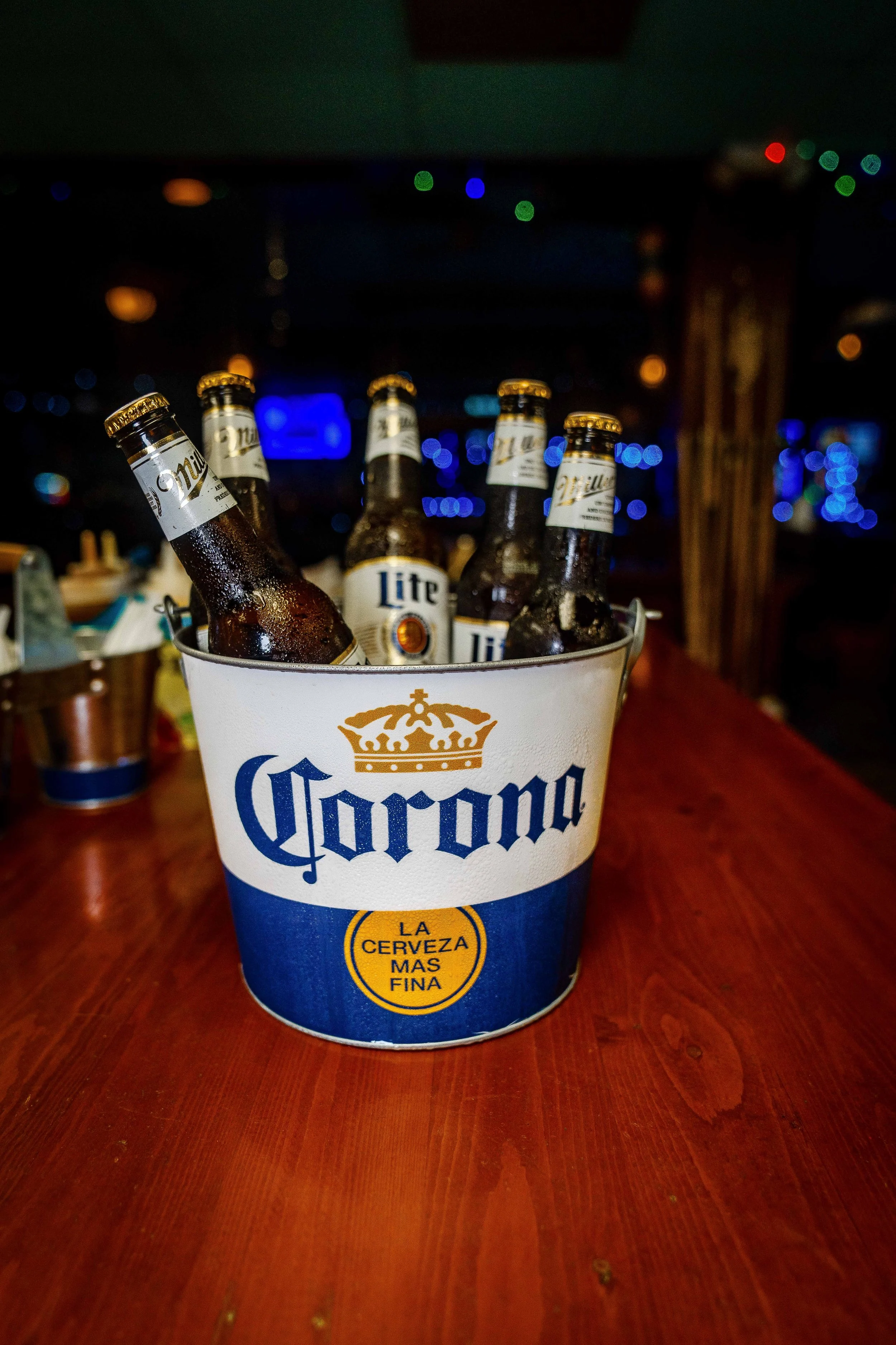 A bucket of six Corona beer bottles on a wooden table in a dimly lit bar or restaurant.