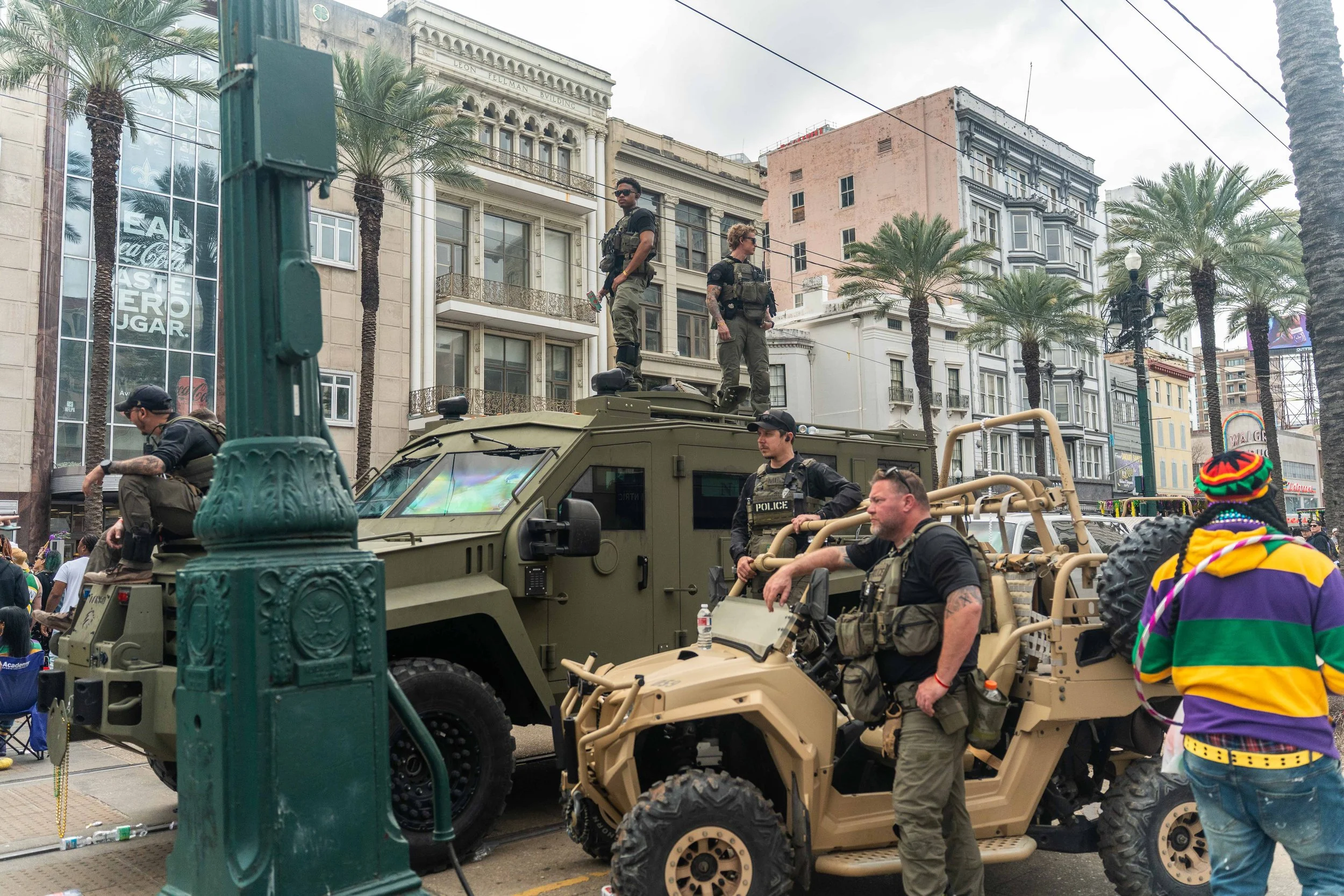 7.0 during Mardi Gras Several police officers in tactical gear on and around military vehicles in a city street, with tall buildings, palm trees, and some people in the background.