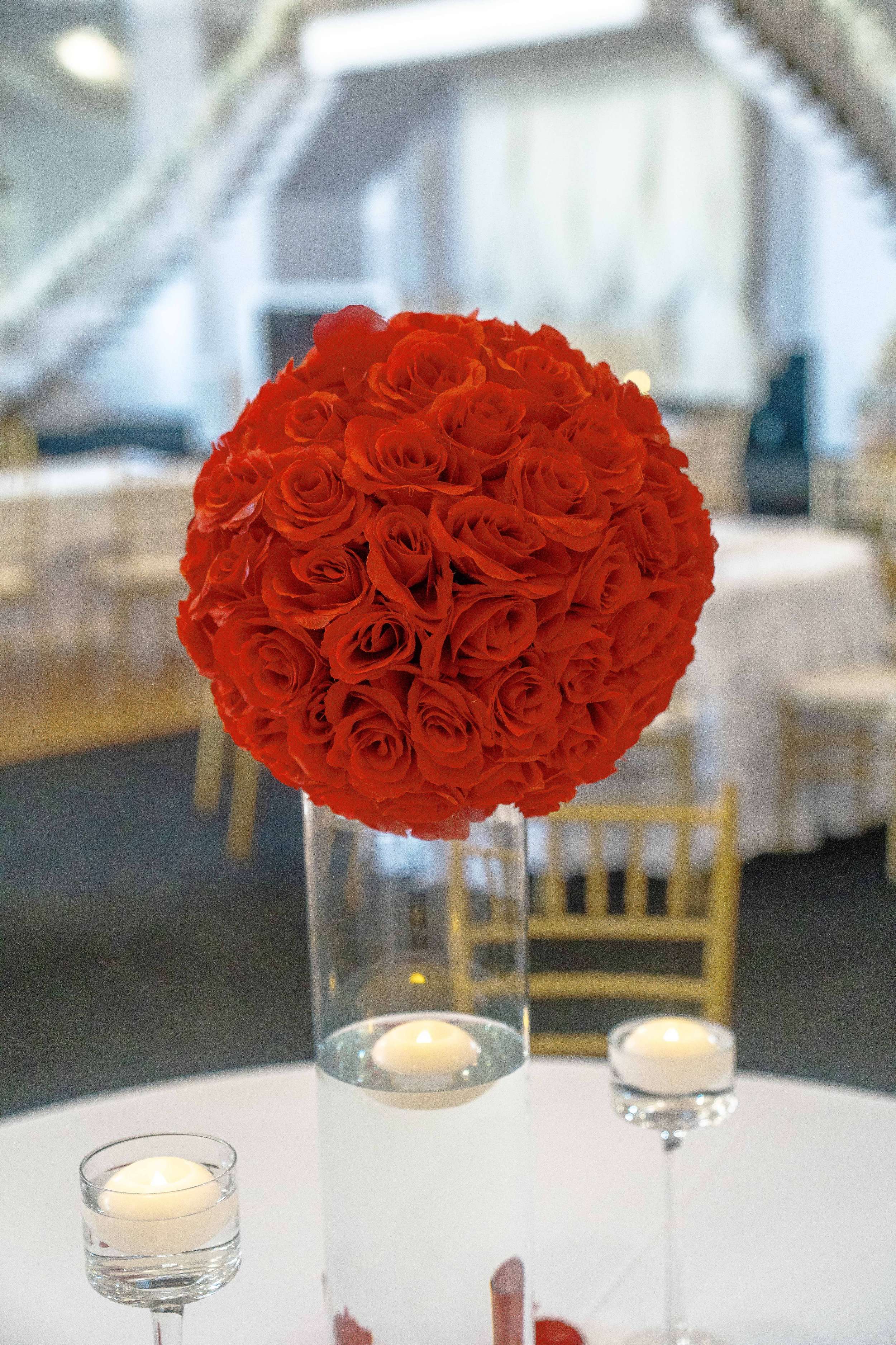 A floral centerpiece made of orange roses is displayed in a tall, clear glass vase. The backdrop appears to be an elegant indoor setting with a blurred background and yellow chairs, illuminated by natural light from large windows. Small tea light candles in glass holders are placed around the floral arrangement on the table.