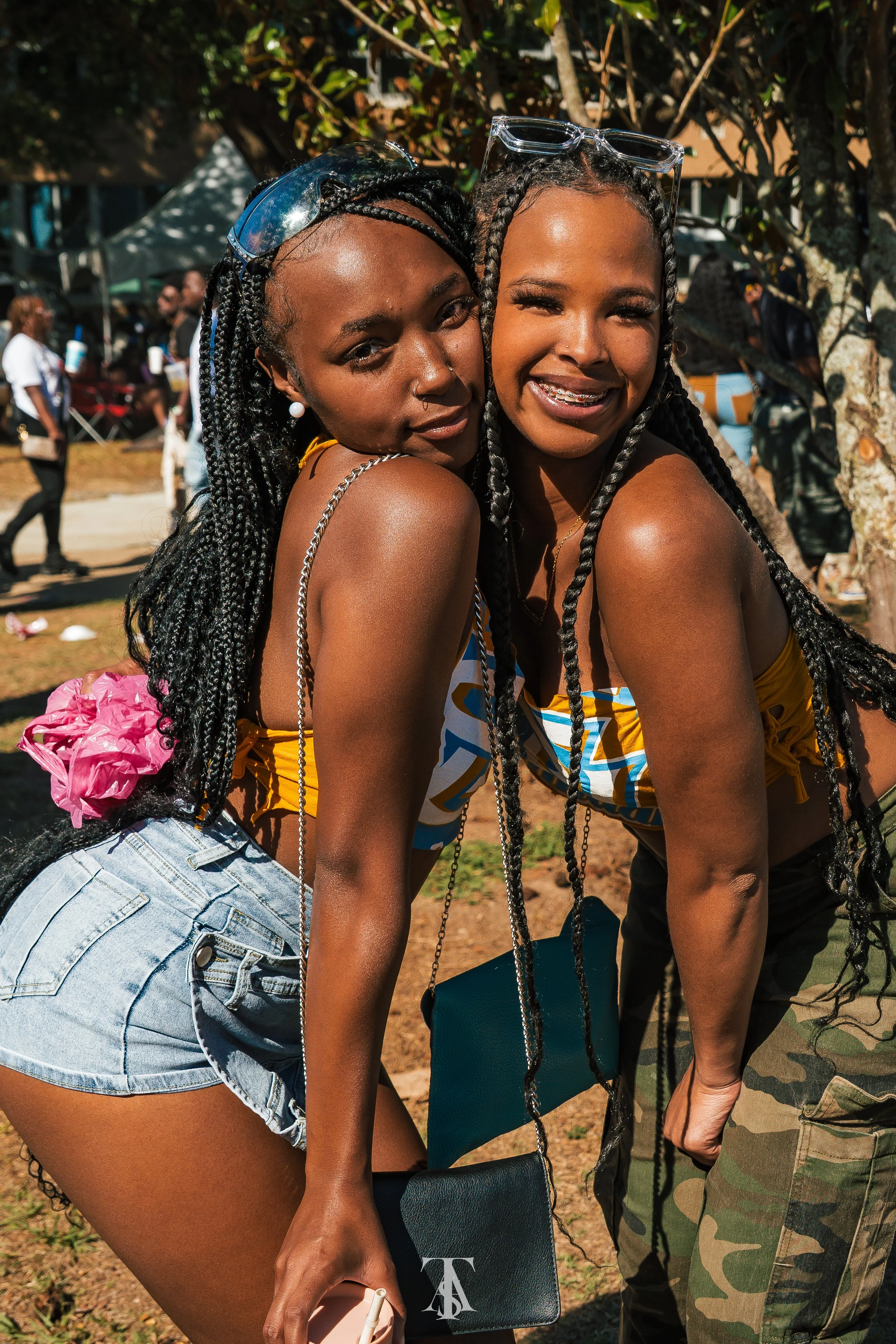 Two women with braided hair and wearing summer clothing, standing close together outdoors at a festival, smiling at the camera.