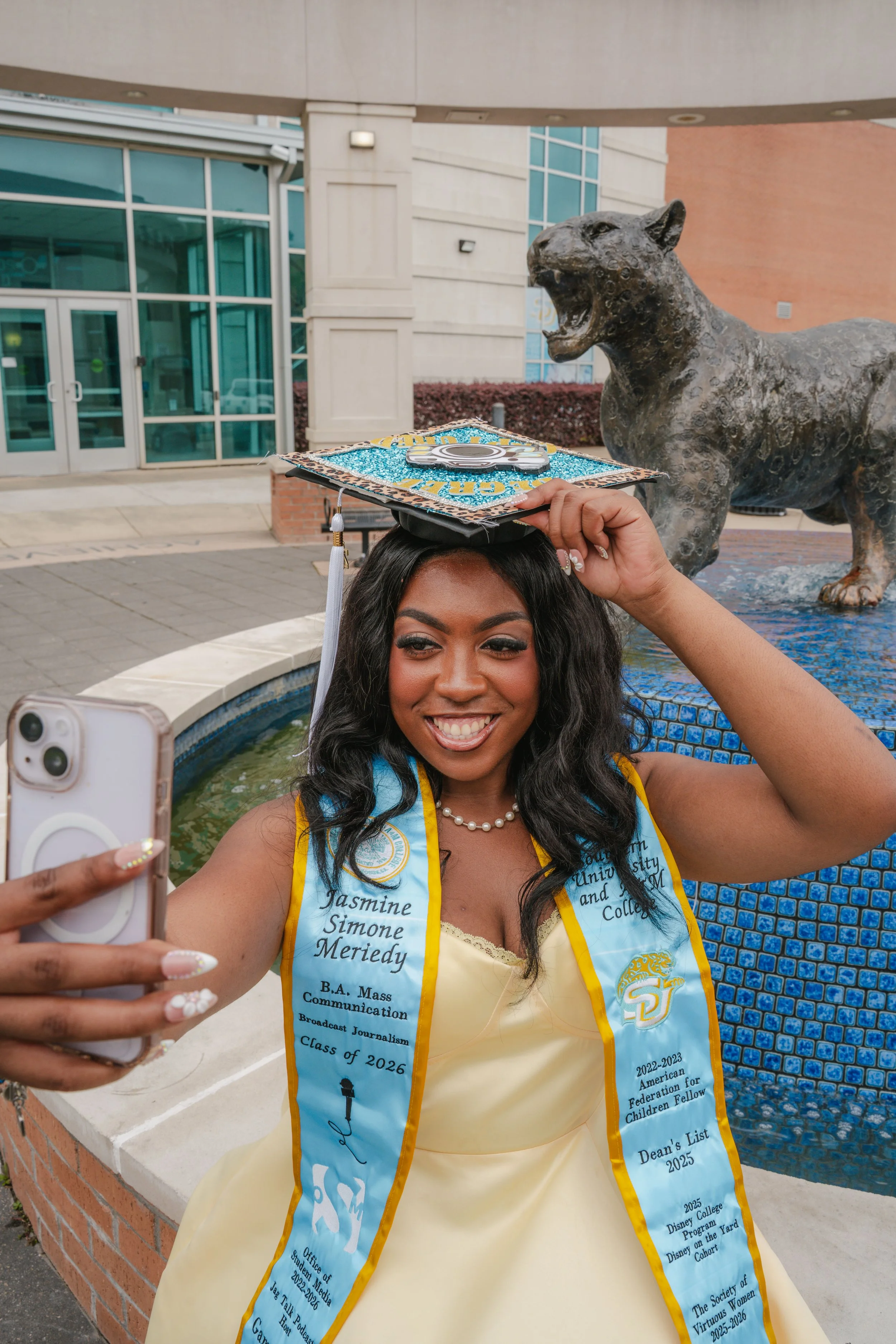 A young woman with long dark hair, wearing a yellow dress and a light blue graduation stole, is smiling and taking a selfie. She is holding her graduation cap with her left hand. Behind her is a fountain with a lion sculpture and a modern building wi