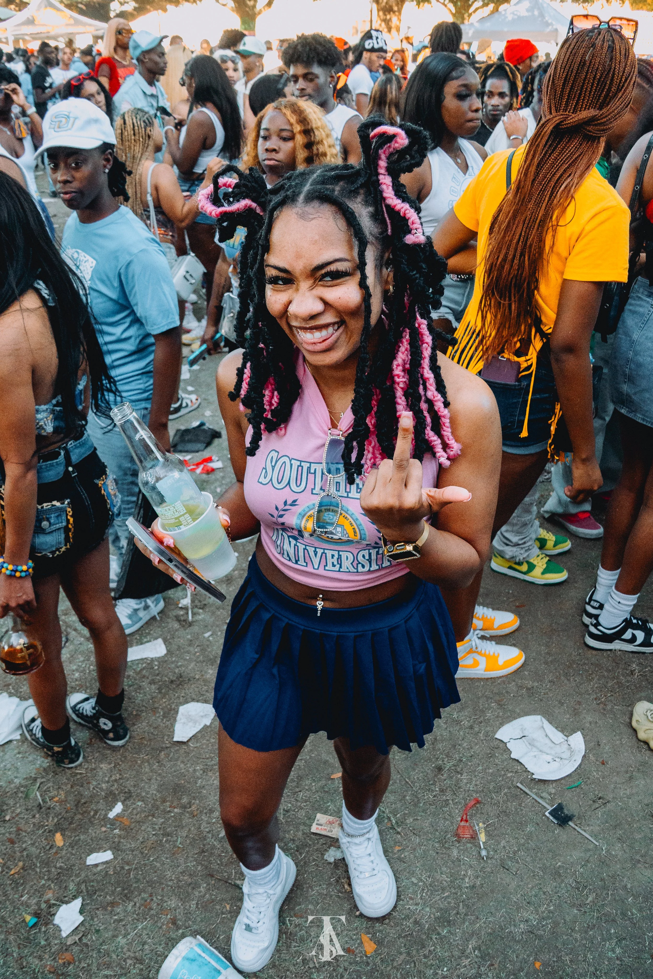 Young woman with colorful braided hair smiling and making a rude hand gesture at a crowded outdoor event.