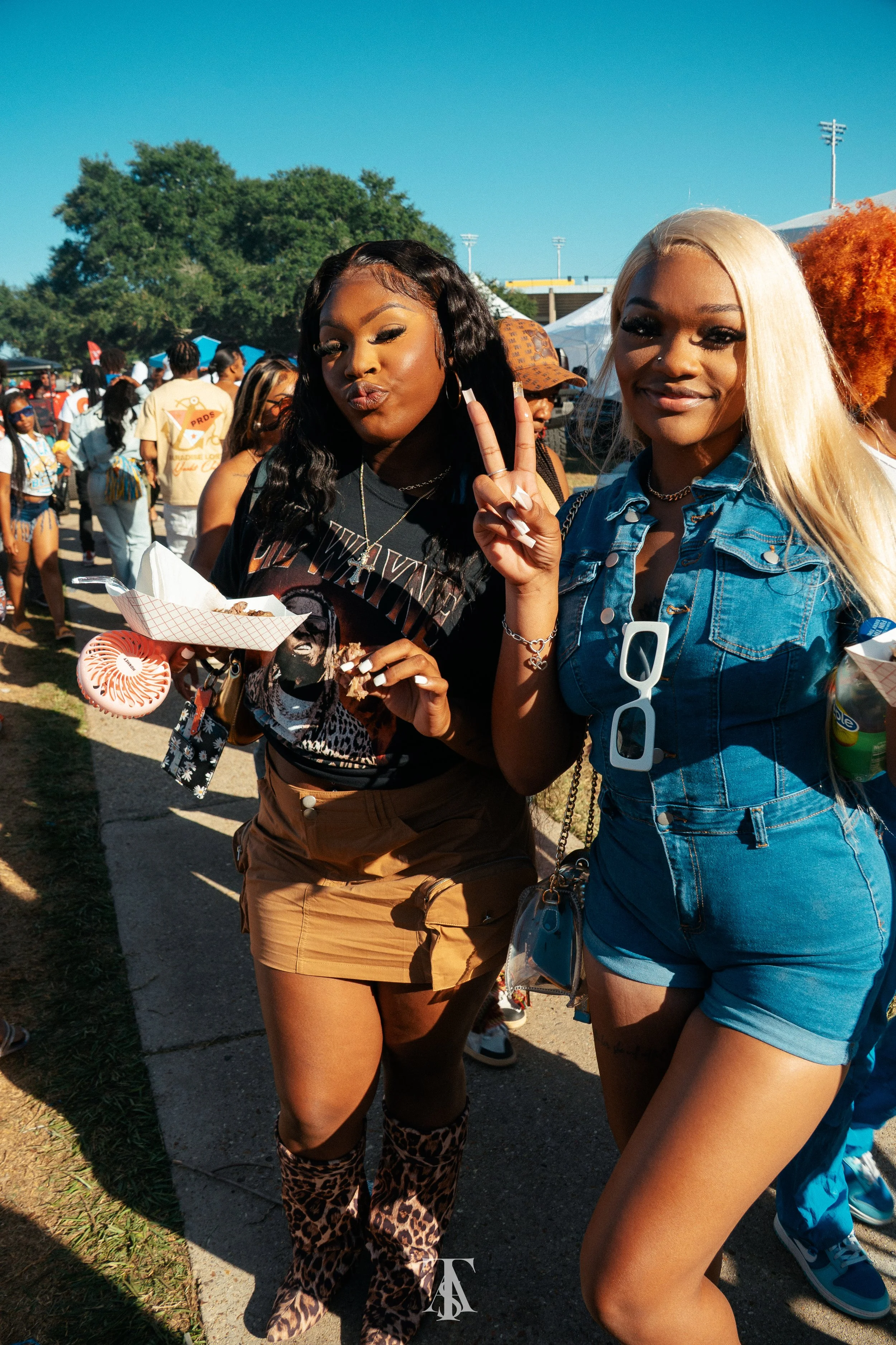Two young women at an outdoor event, one making a peace sign and the other holding food.