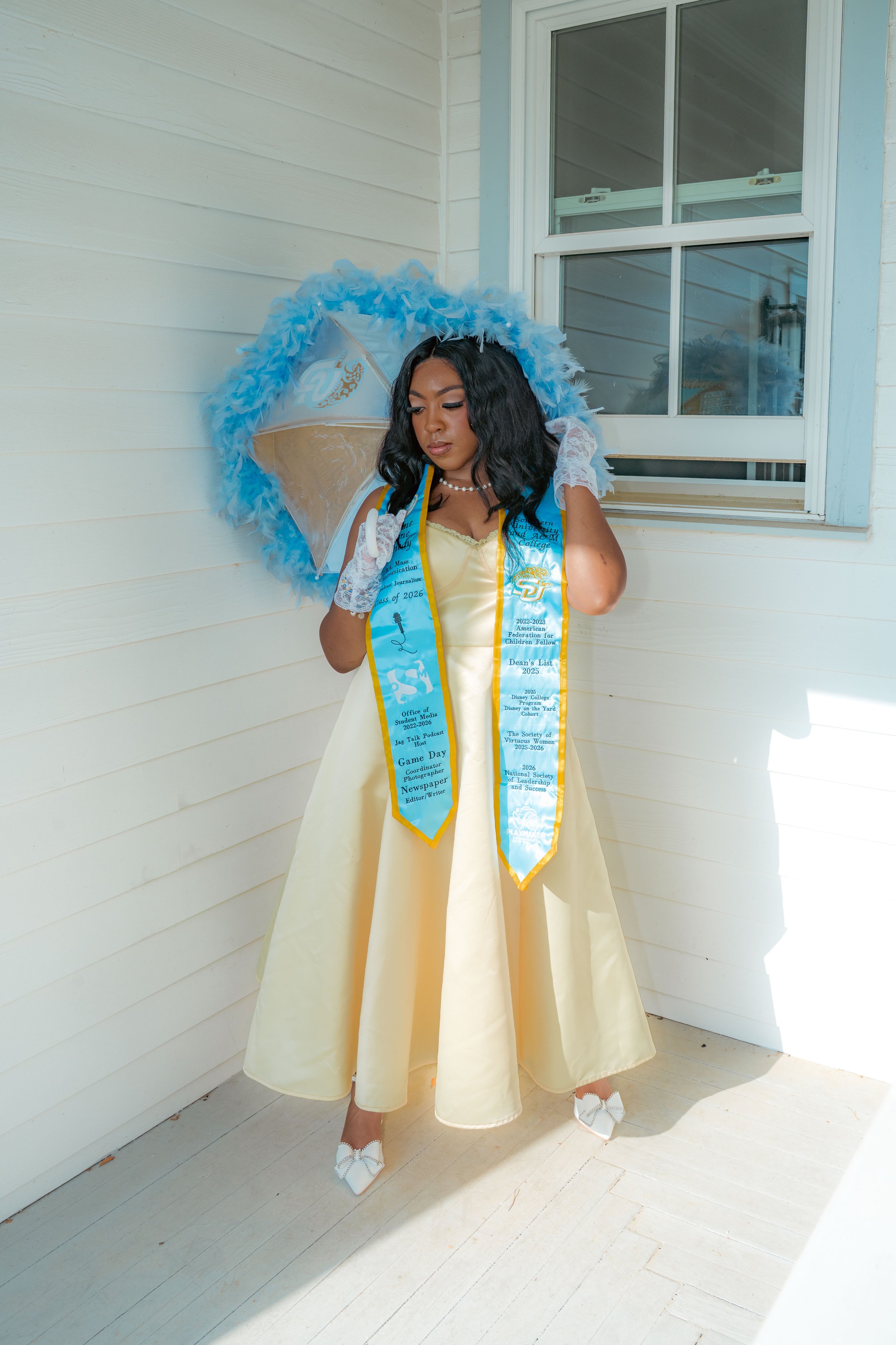 A young woman in a yellow gown standing outside on a porch with cream-colored wooden siding. She wears white gloves, white high heels, and a blue sash with gold trim and text. She holds a clear umbrella decorated with blue feathers, with a blue feath