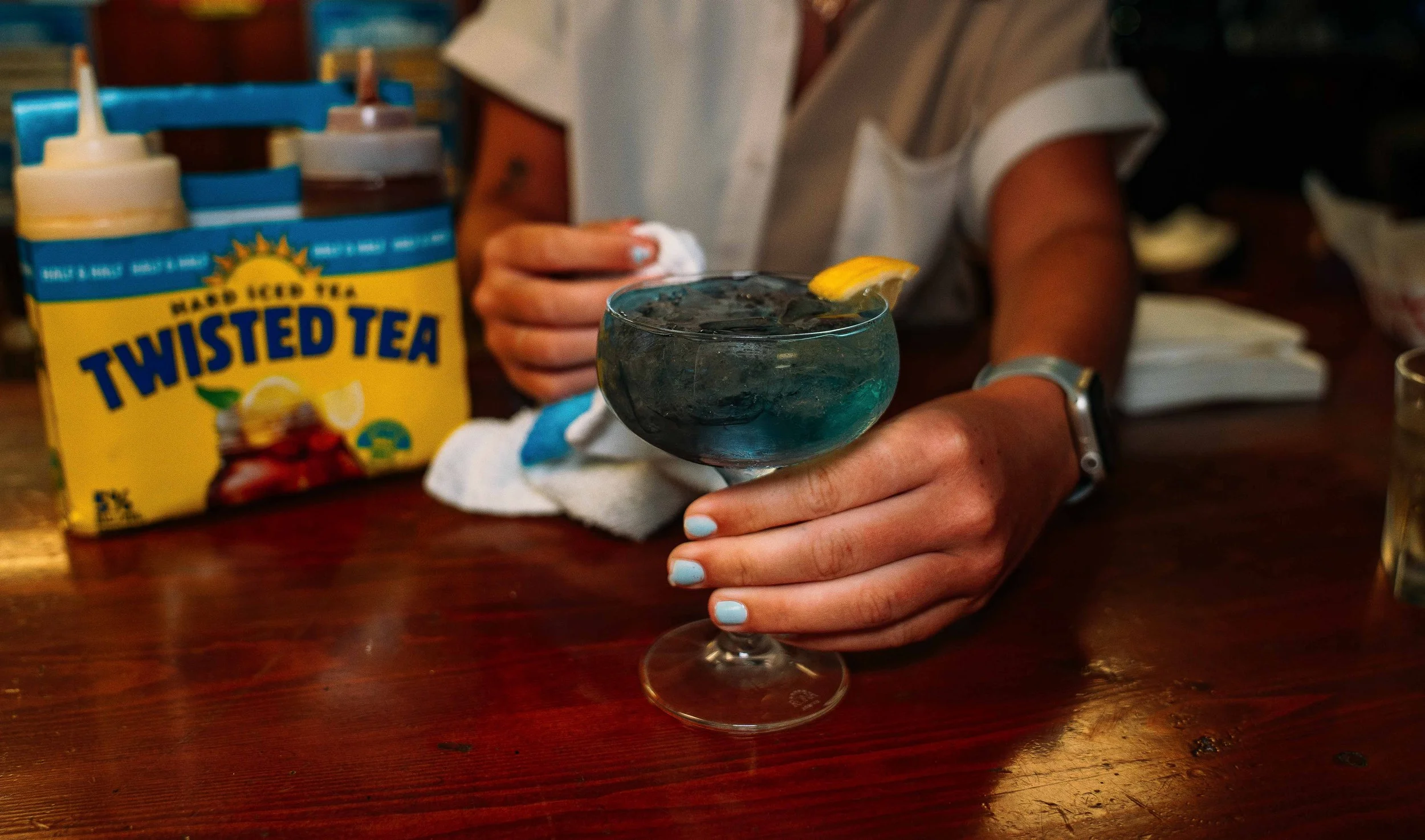 Person holding a cocktail glass with a lemon wedge garnish, behind a package of Twisted Tea and a hand towel on a wooden table.