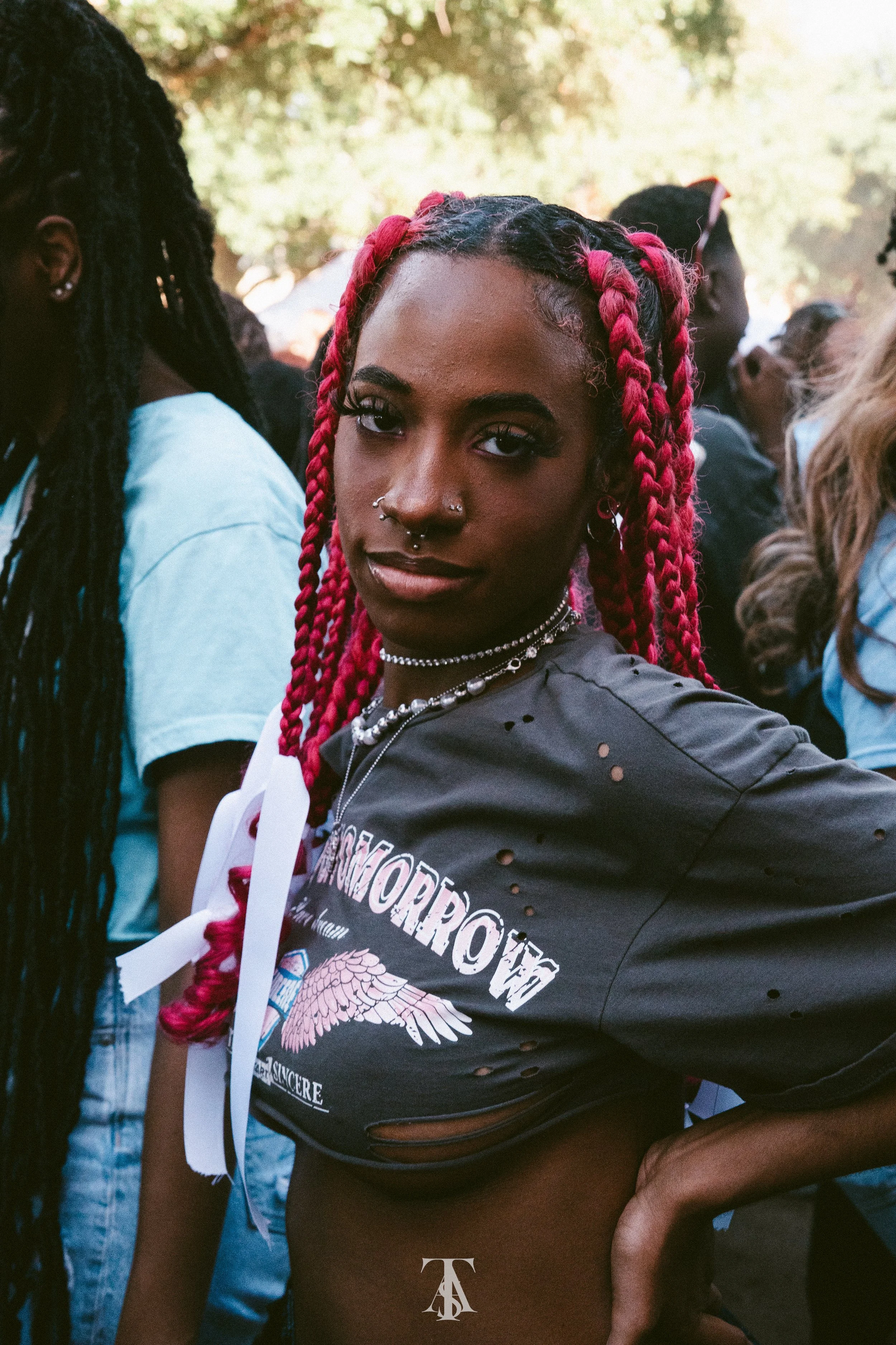 A young woman with bright pink braids, piercings, and jewelry looks confidently into the camera at an outdoor gathering with trees in the background.