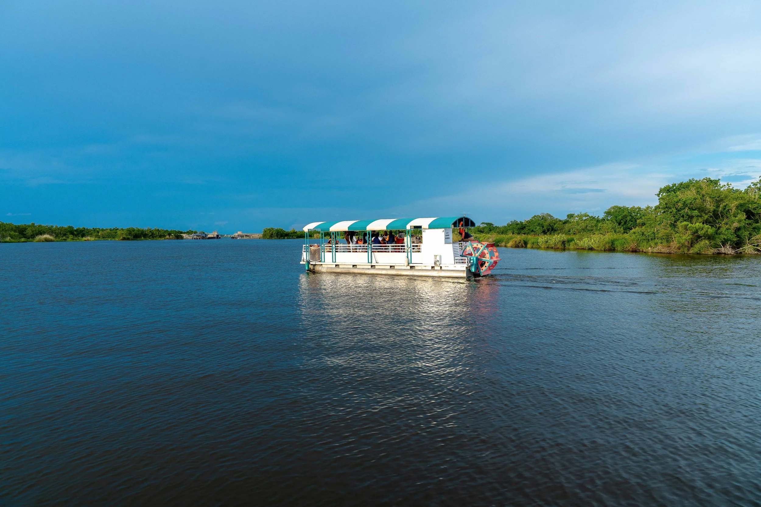 A boat with a canopy carrying people on a calm river, surrounded by green trees and under a cloudy sky.
