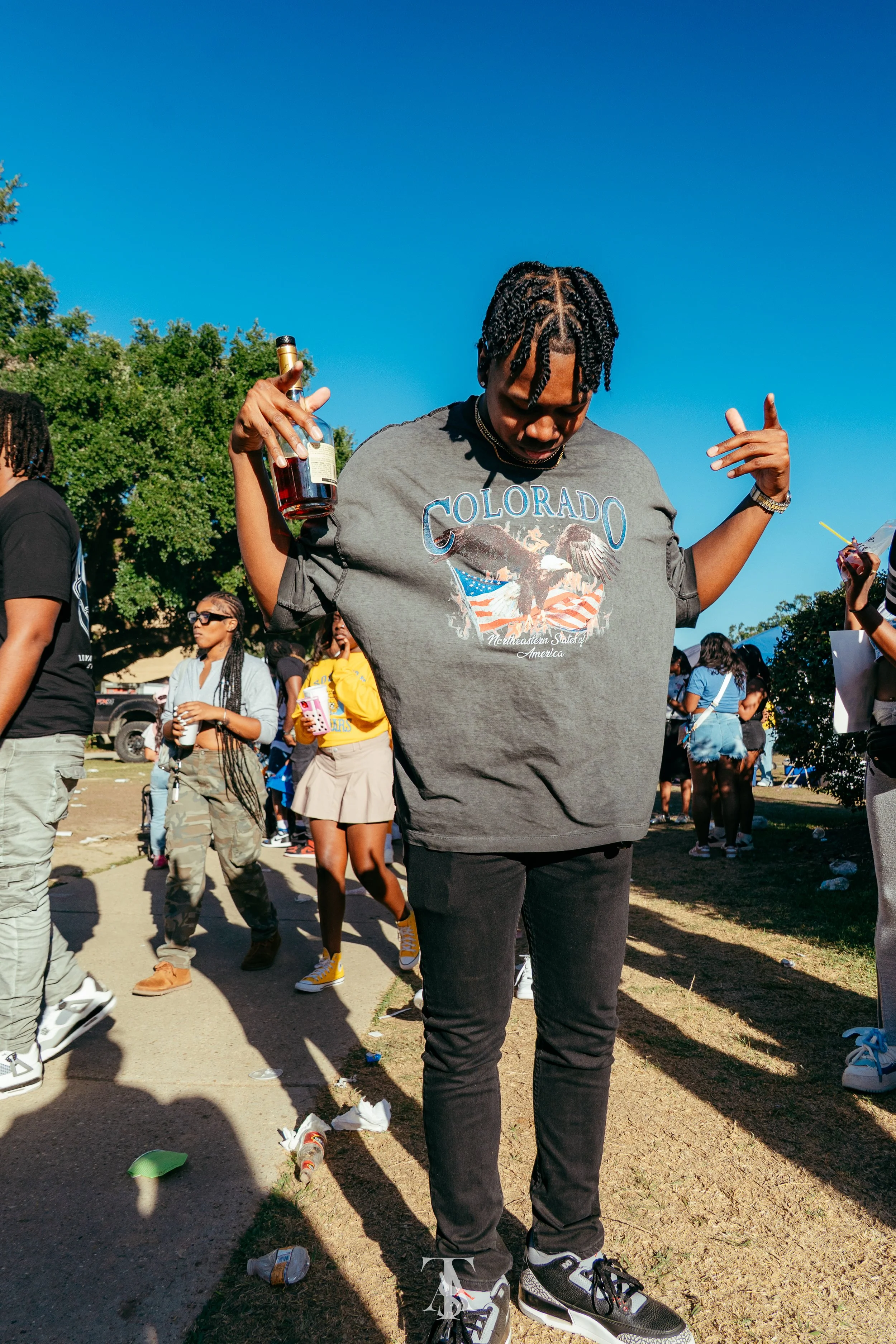 A young man with twists hairstyle holding a bottle of alcohol while dancing at an outdoor event, surrounded by other people on a sunny day.
