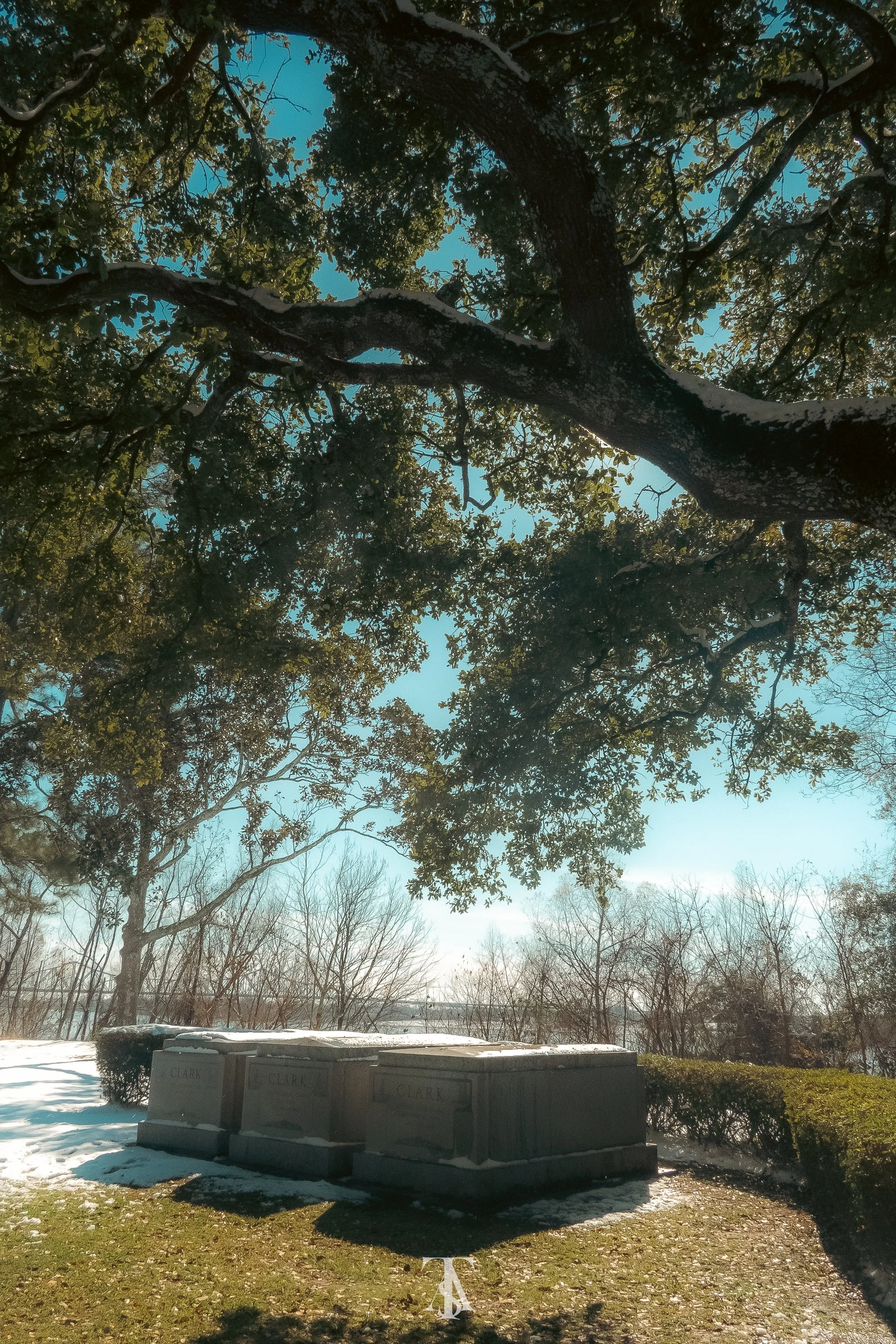 A park scene with large trees, a stone monument labeled "Clark," snow on the ground and bushes, and a clear blue sky.