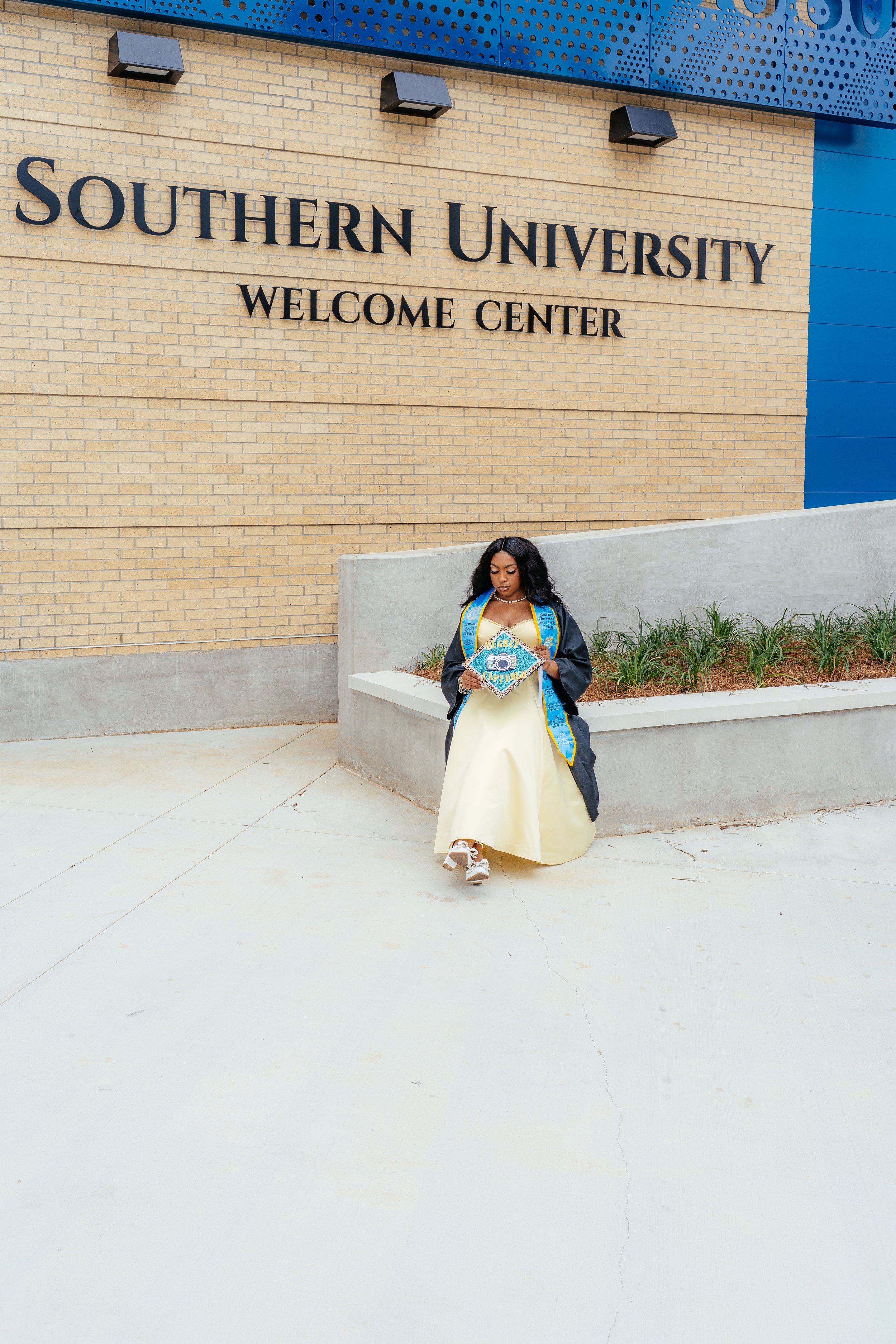 A woman in a yellow dress and black graduation gown stands outside Southern University Welcome Center, holding her diploma and walking on a concrete pathway.