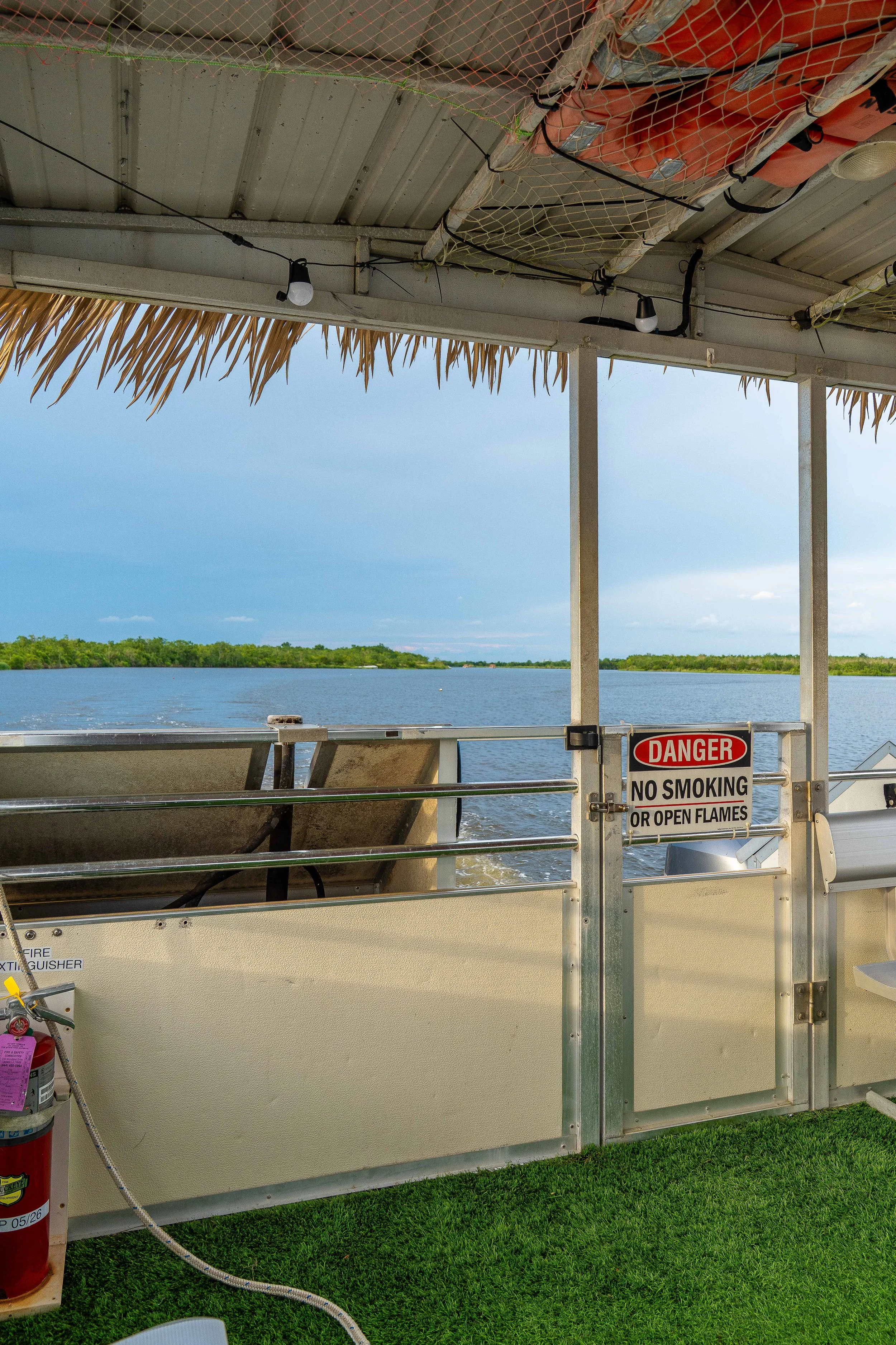 View of a dock or boat area overlooking water with trees in the distance. The foreground features a metal railing, a fire extinguisher, and a warning sign that says 'DANGER NO SMOKING OR OPEN FLAMES.' The top part of the image shows a thatched roof, 