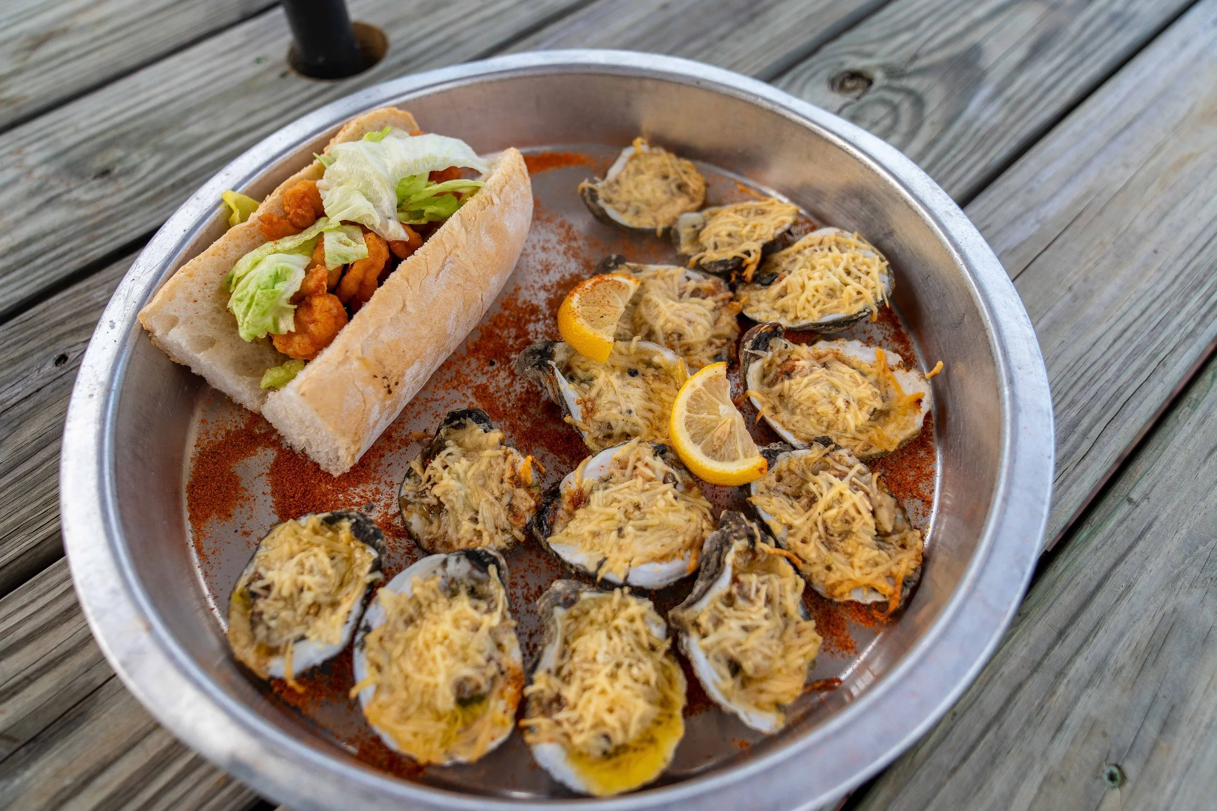 A round metal tray with grilled oysters topped with cheese and lemon slices, and a steamed bun sandwich filled with lettuce and fried chicken, placed on a wooden surface.