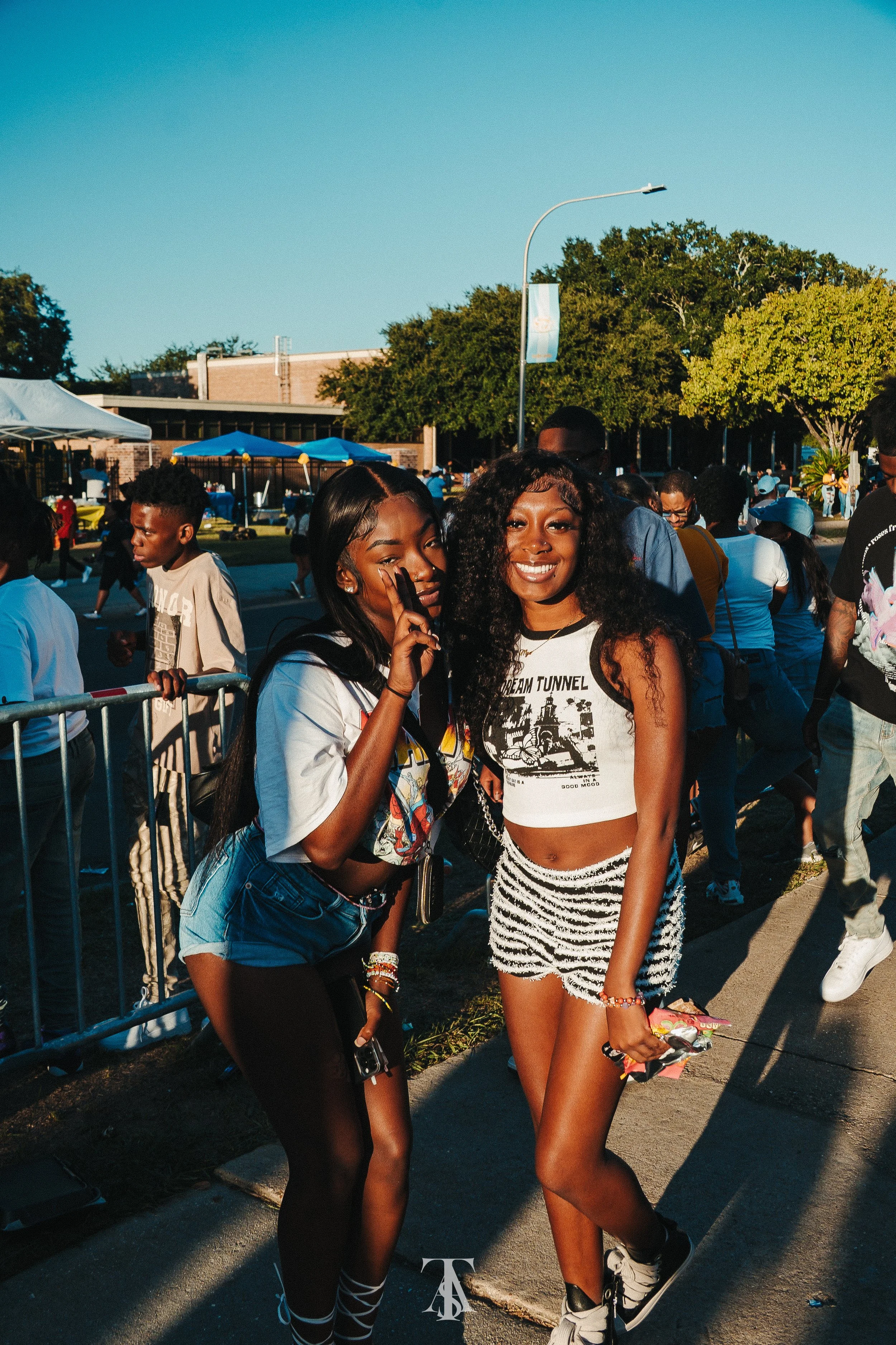 Two young women posing together outdoors at a crowded event with tents and trees in the background, smiling and making peace signs.