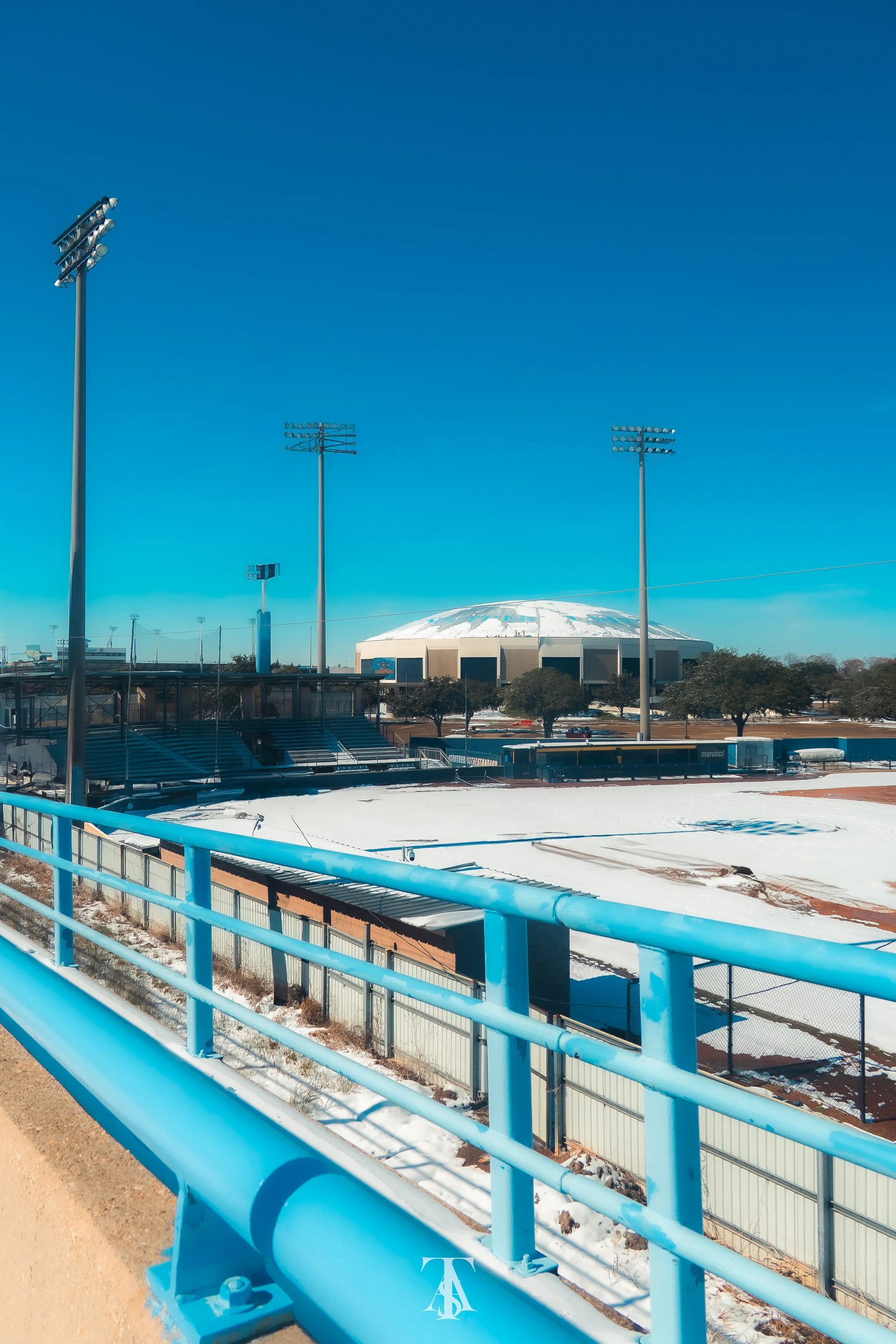 A stadium with snow on the field, tall light poles, and a domed roof in the background, viewed from a blue railing along a walkway.