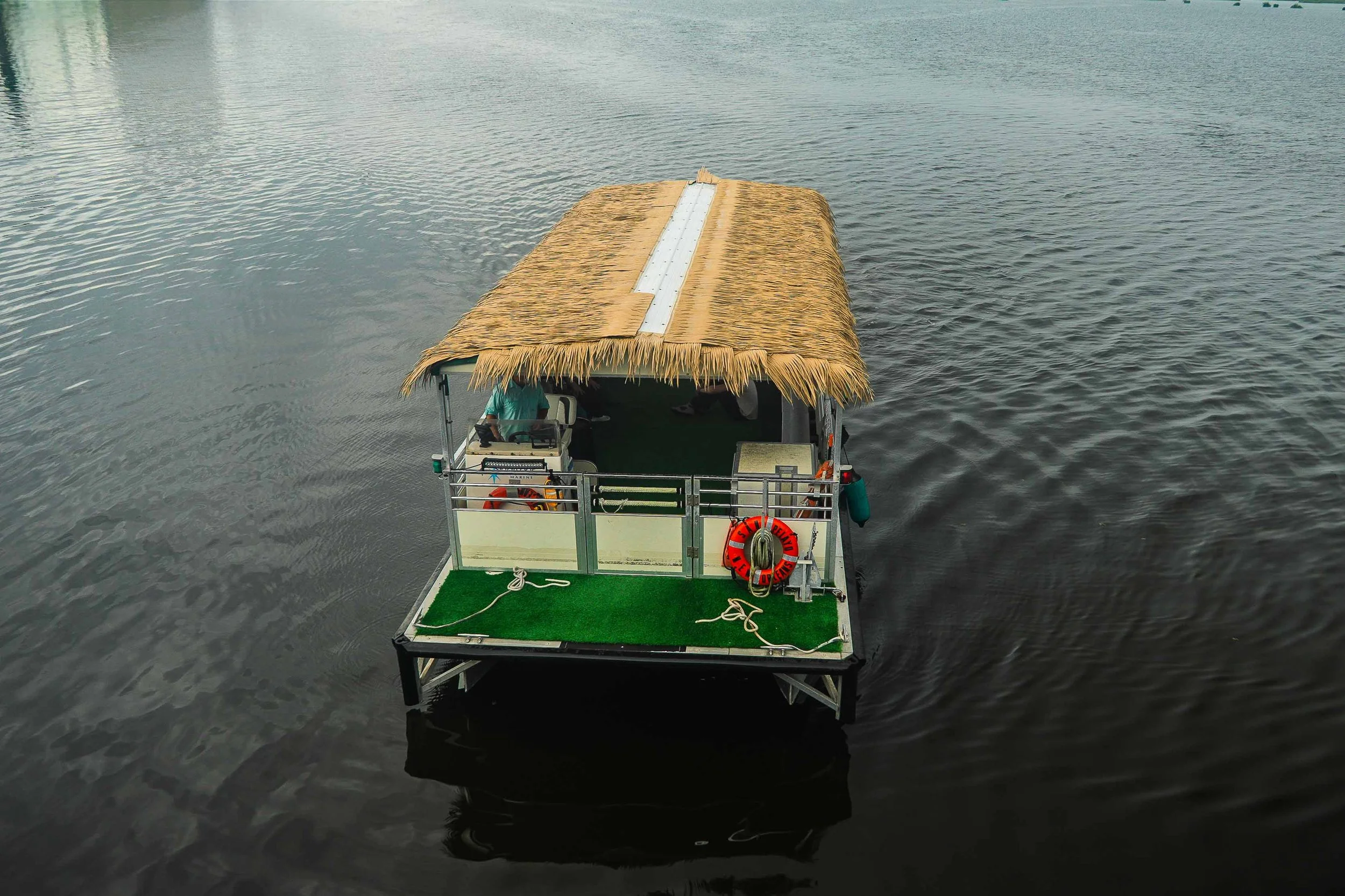 A boat with a thatched roof and a green artificial turf deck floating on dark water, with a person standing at the helm.