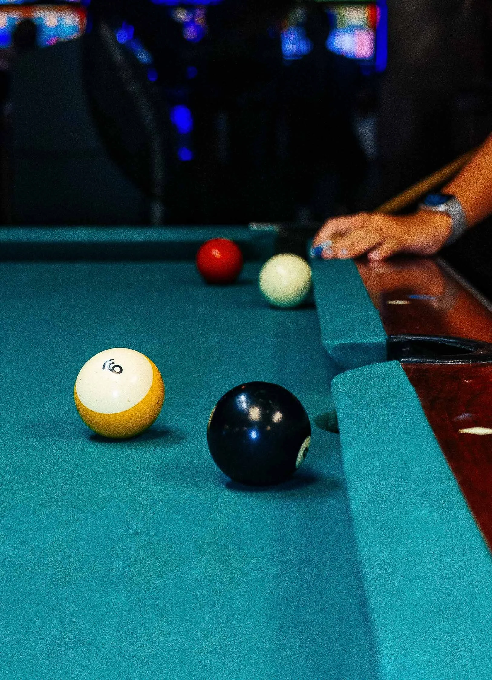 Close-up of a billiard table with a white, yellow, red, and black balls, and a person's right hand aiming with a cue stick.