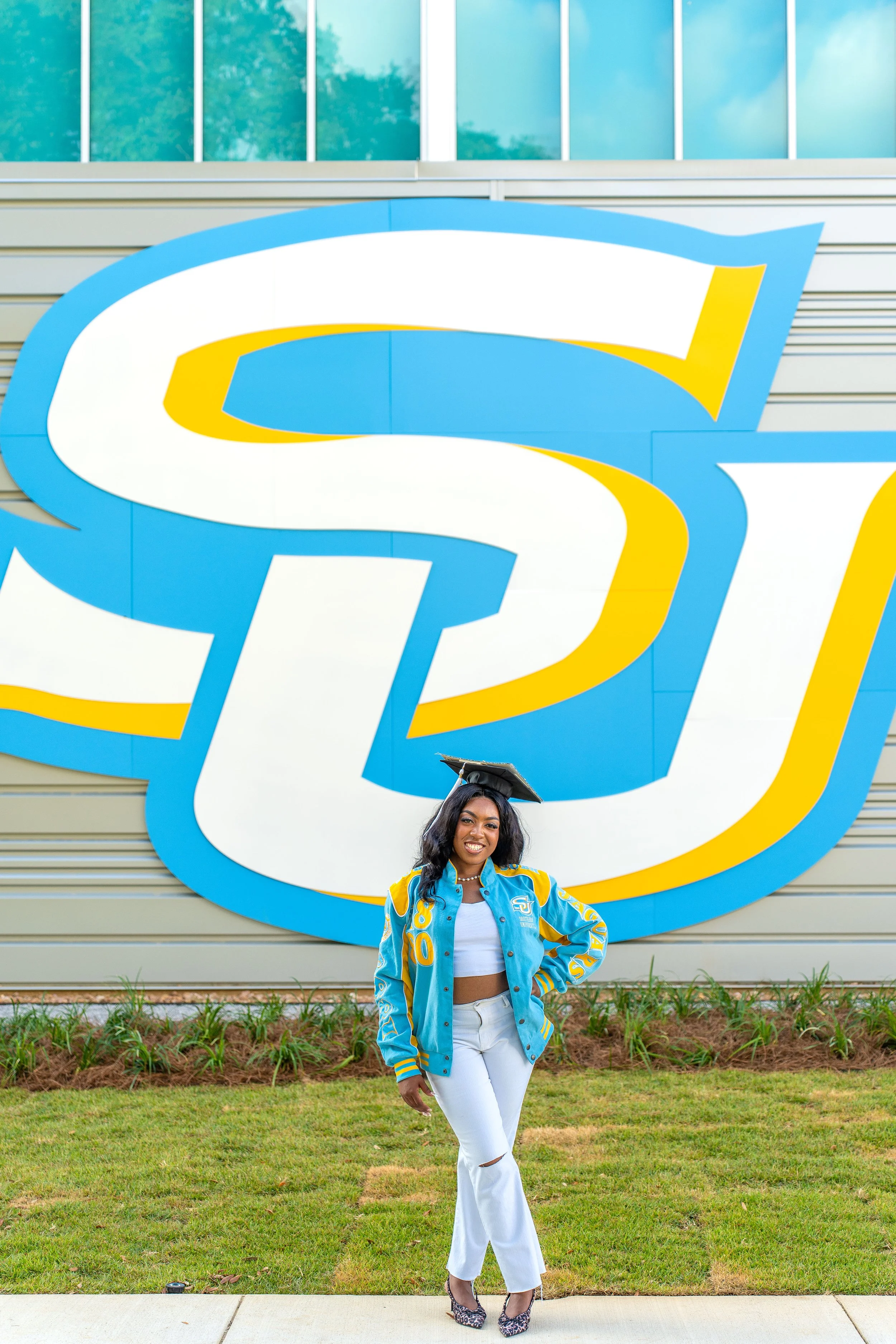 A young woman in a blue and yellow jacket and white pants stands outside in front of a large retro sign with a stylized 'S' and 'U' logo. She is smiling, and a graduation cap is on her head. The background includes a building with large blue-tinted w