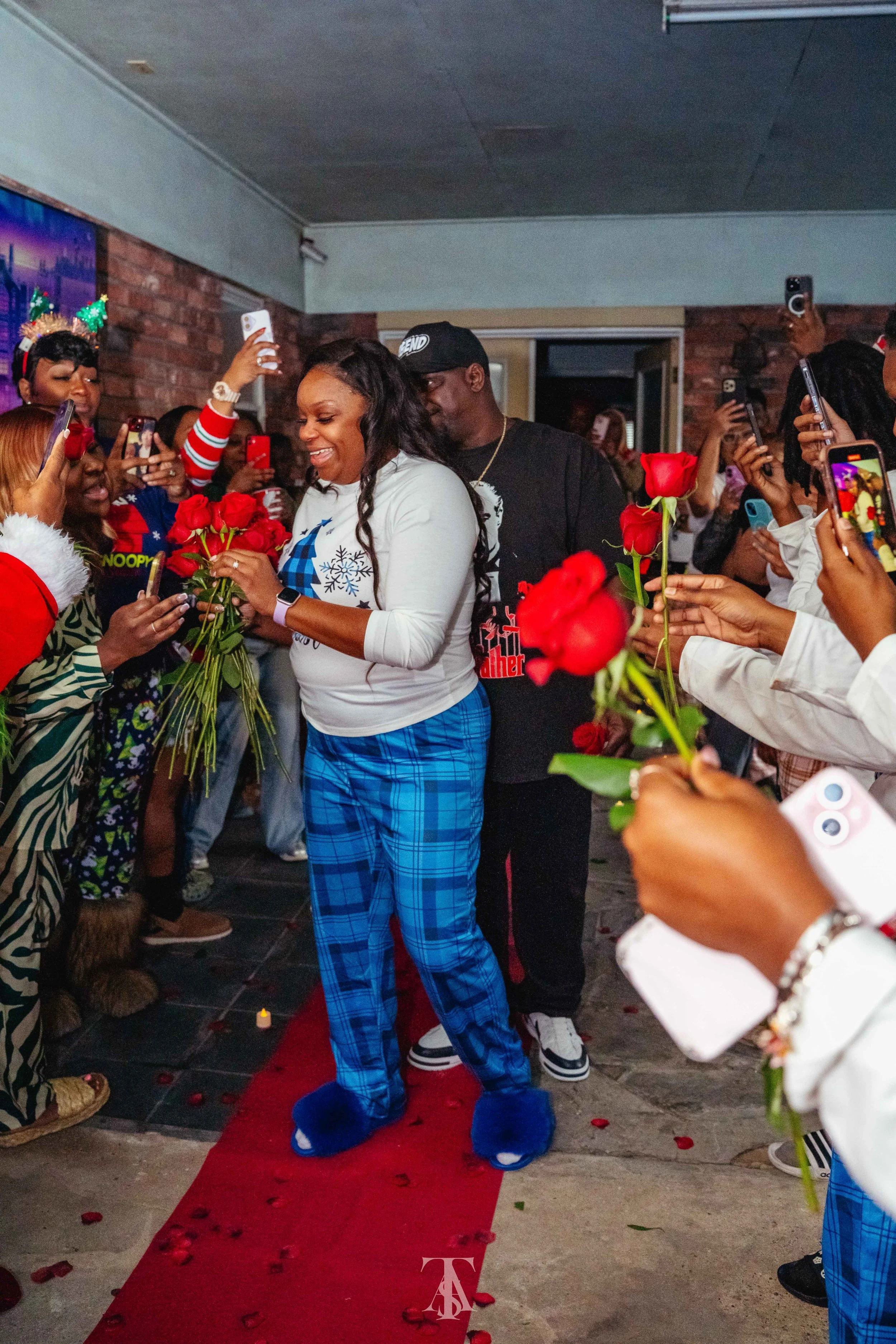 A woman in blue plaid pajama pants and fluffy blue slippers is standing on a red carpet, holding a bouquet of red roses, surrounded by people taking photos and holding roses, at an indoor event decorated with rose petals for a celebration.