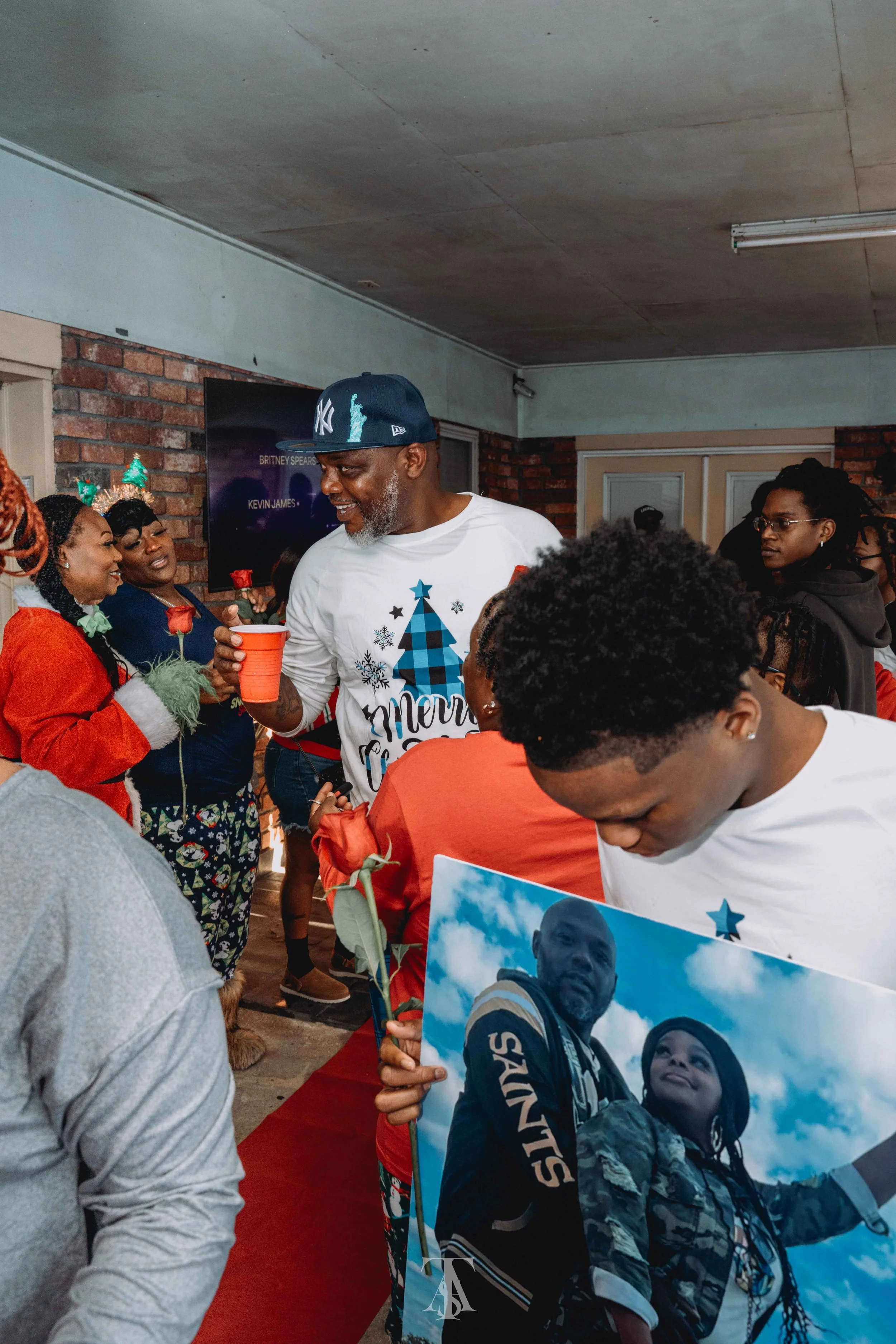 People gathering indoors, engaging in conversation, some holding roses and posters, with a brick wall and a TV screen displaying names in the background.
