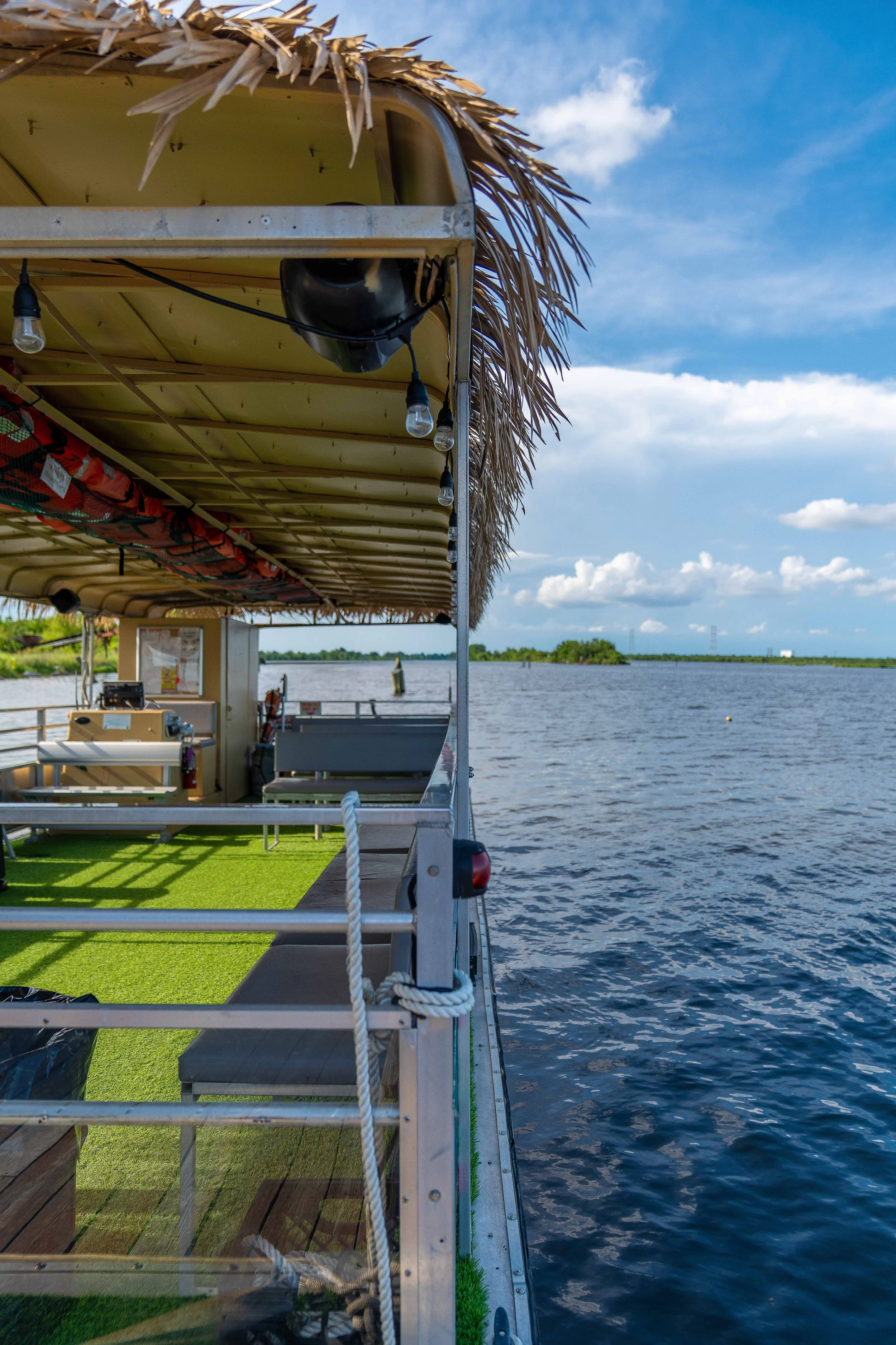 Side view of a boat with a green artificial turf deck, that is floating on a body of water, with a thatched roof and hanging light bulbs, under a partly cloudy sky.