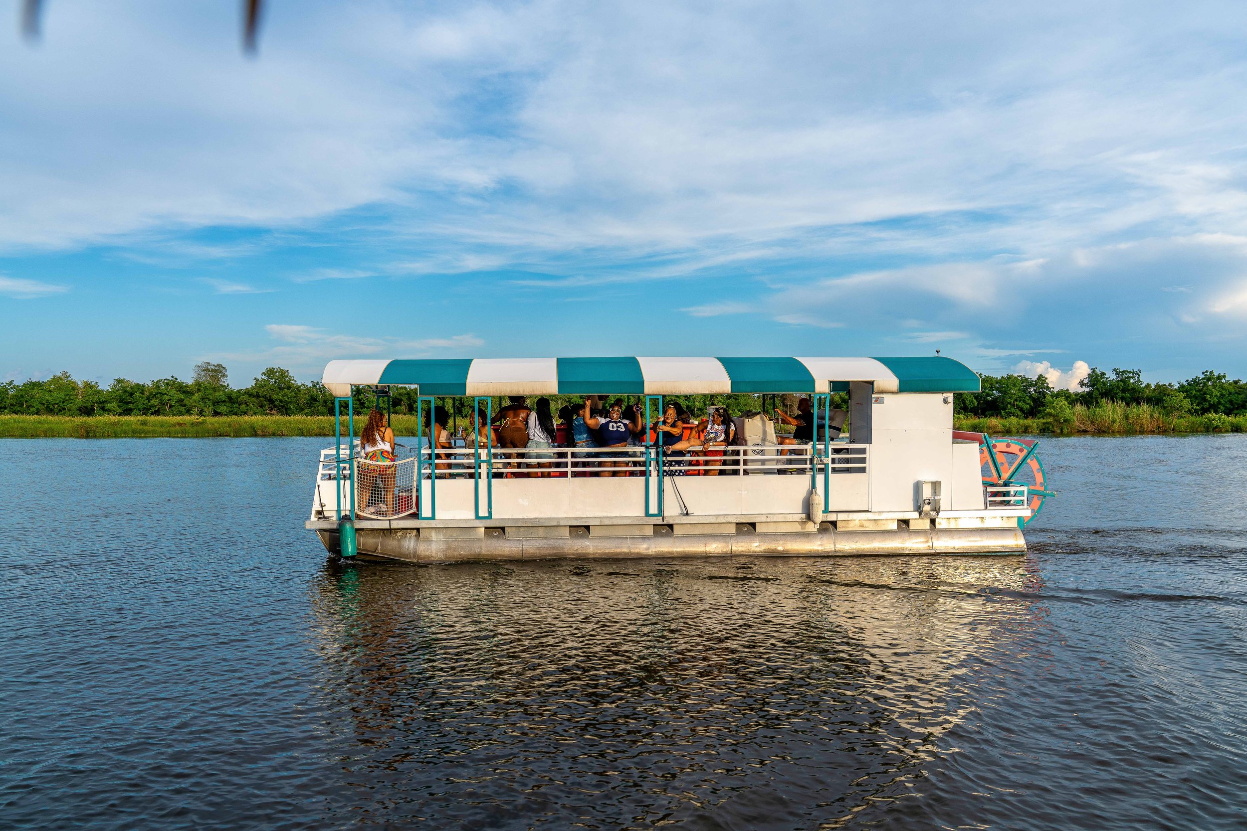 People enjoying a boat ride on a calm river under a partly cloudy sky, surrounded by lush green trees.