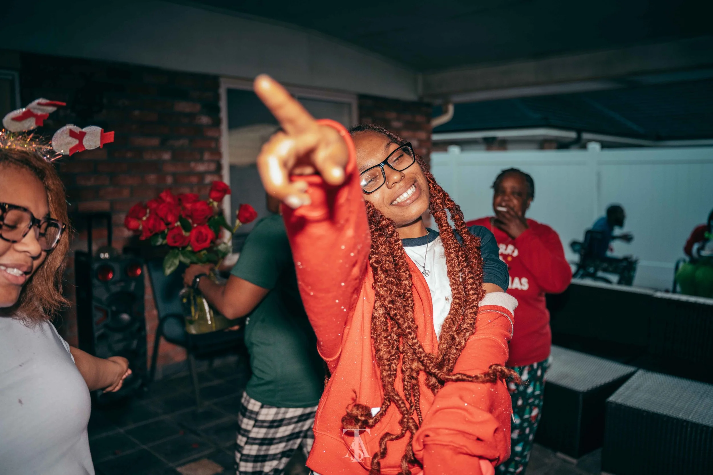 A woman with glasses and dreadlocks smiling and pointing at the camera, wearing an orange jacket, with others around her celebrating indoors.
