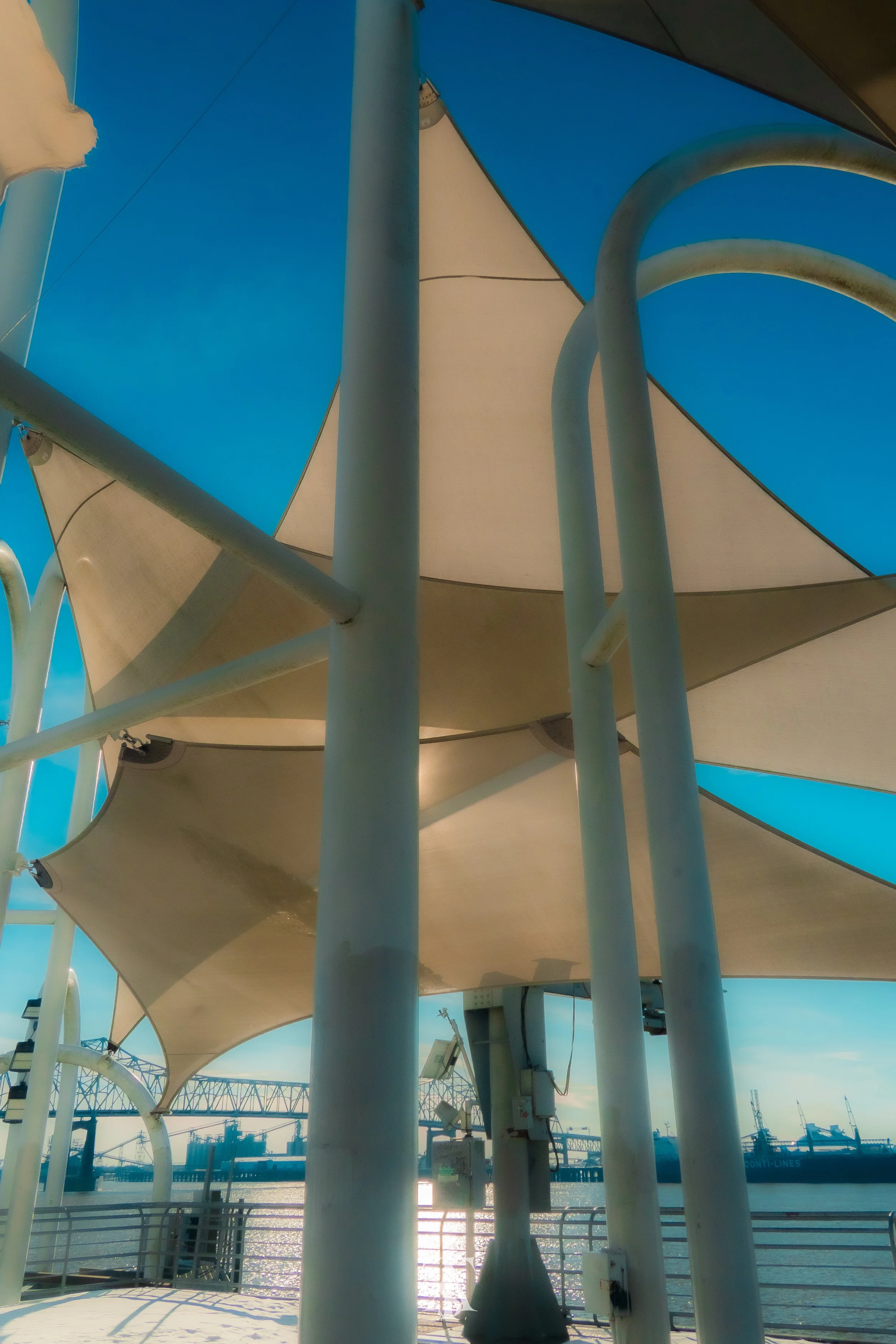View of a waterfront with sail shade structures, metal poles, and weather stations or equipment under a clear blue sky.