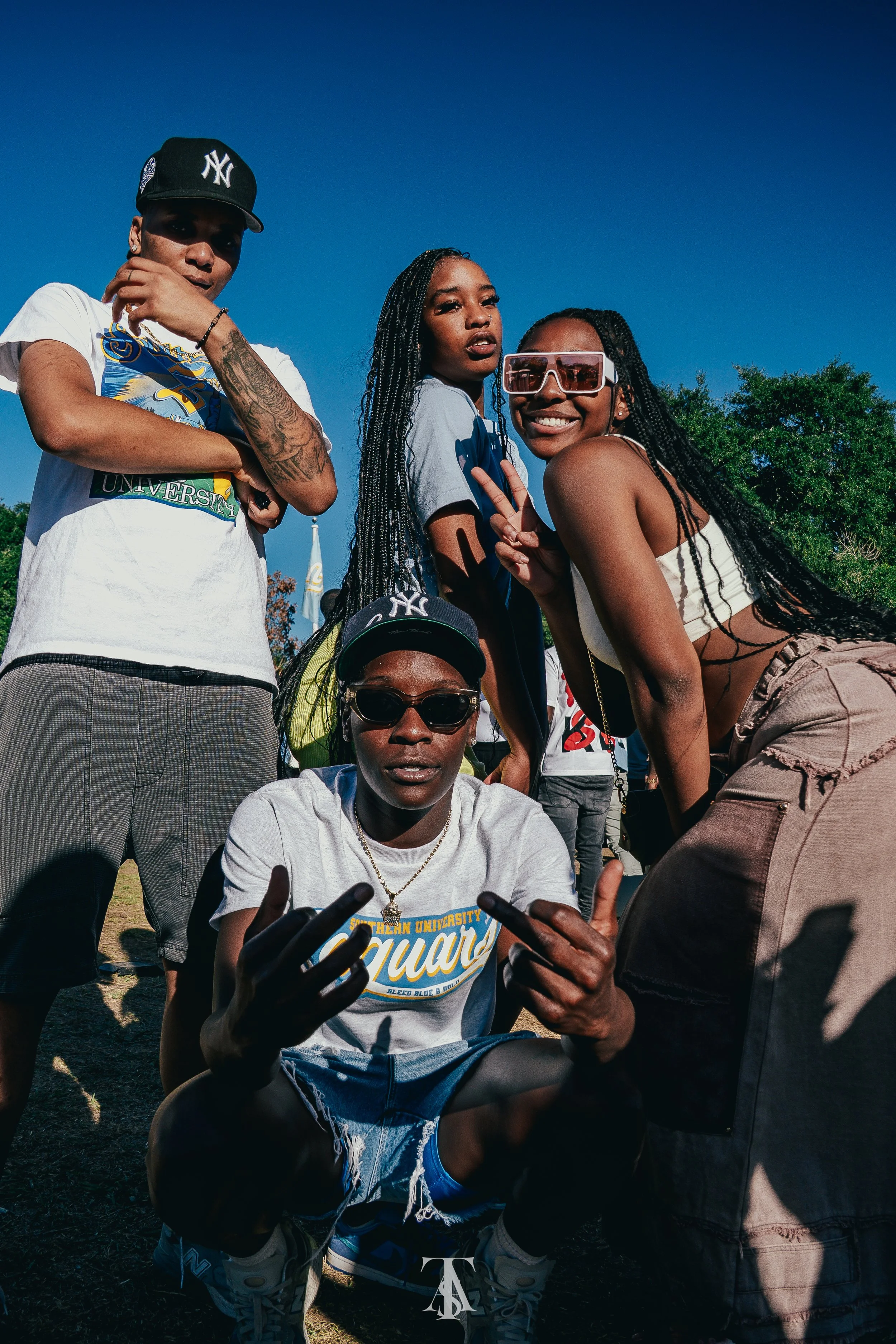Five young adults outdoors under a clear blue sky, posing for a photo. They are dressed casually, with some wearing sunglasses and hats, displaying confident and friendly expressions.