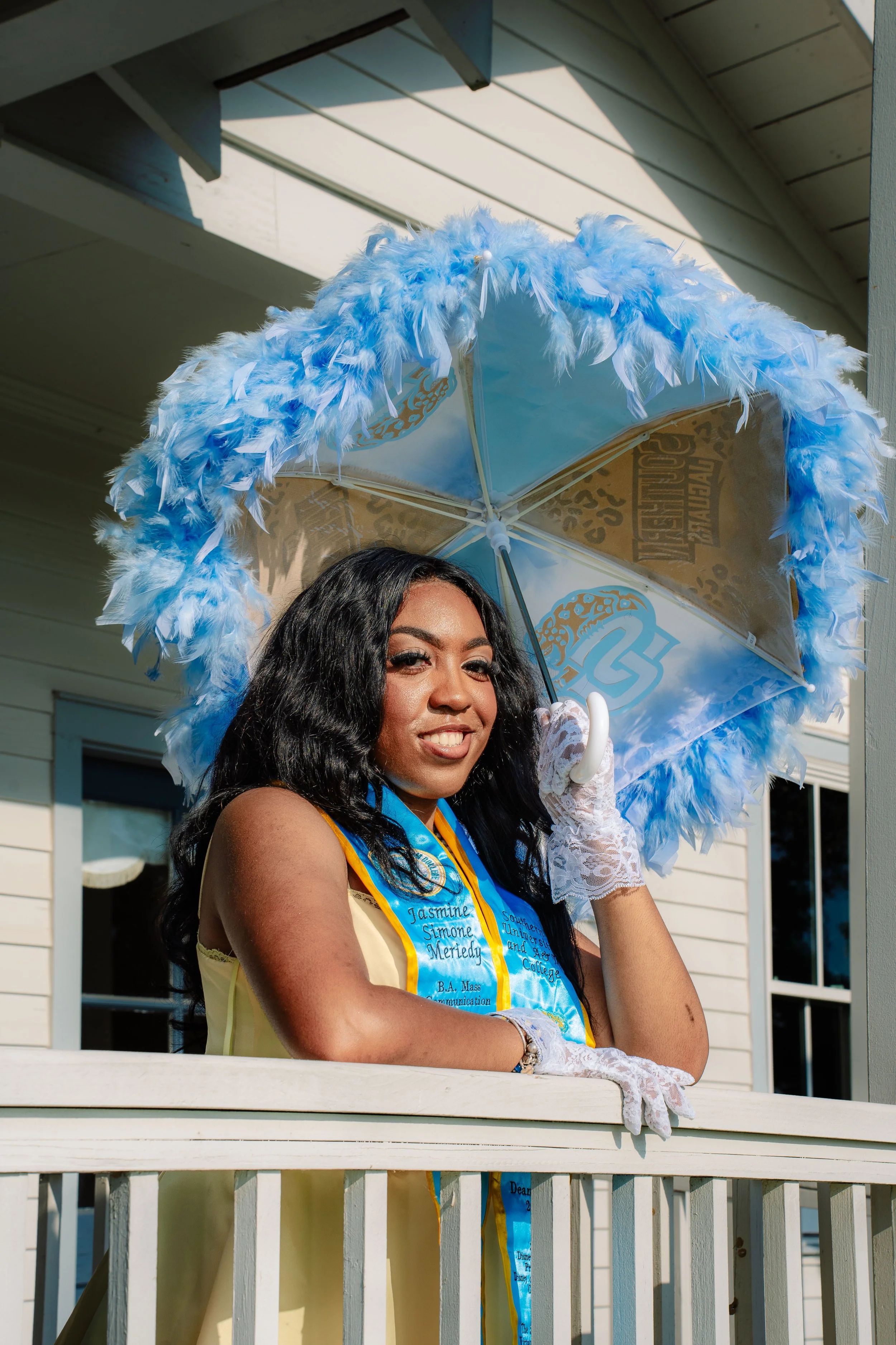 Young woman in yellow dress standing behind a white porch railing, holding a white lace glove and an umbrella decorated with blue feathers, sunlit outdoor setting.
