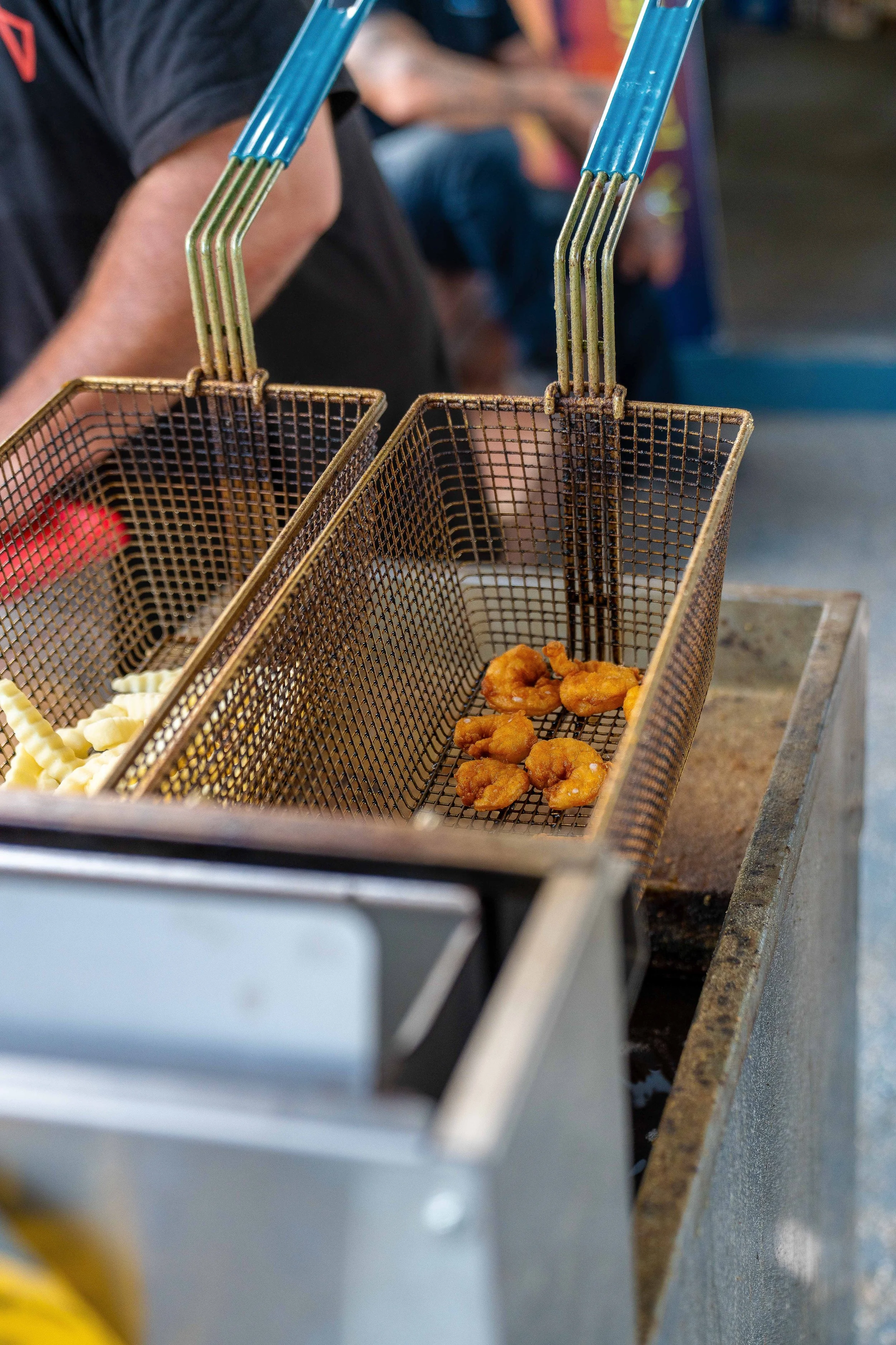 A person frying small pieces of battered food, possibly chicken, in a wire basket over hot oil on a countertop in a commercial kitchen or food stall.