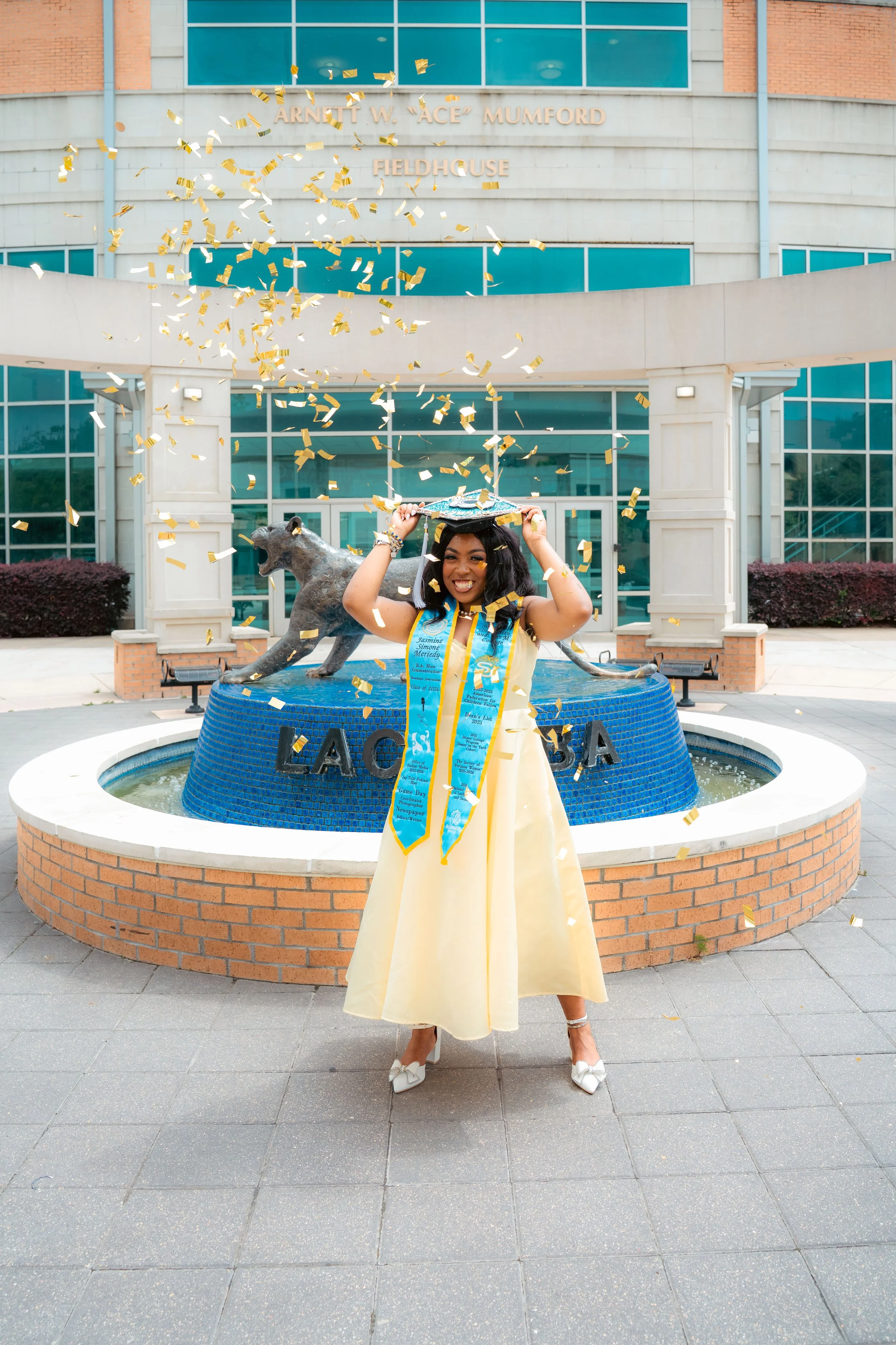 A woman in a yellow dress and white high heels celebrating her graduation in front of a courthouse fountain with a bronze statue of a panther. She is holding her graduation cap and wearing a blue sash. Gold and white confetti are falling around her.