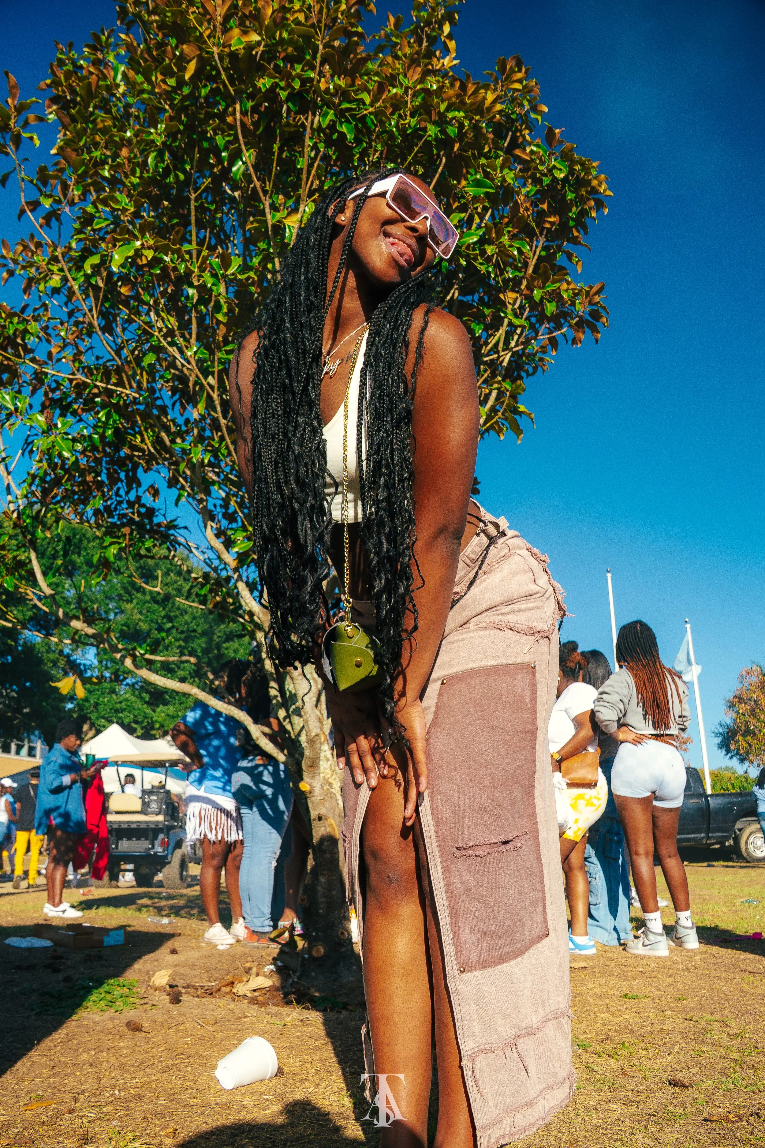 A young woman with long braids wearing sunglasses, a white top, and ripped beige pants, standing outdoors during daytime with a tree and a crowd of people in the background.