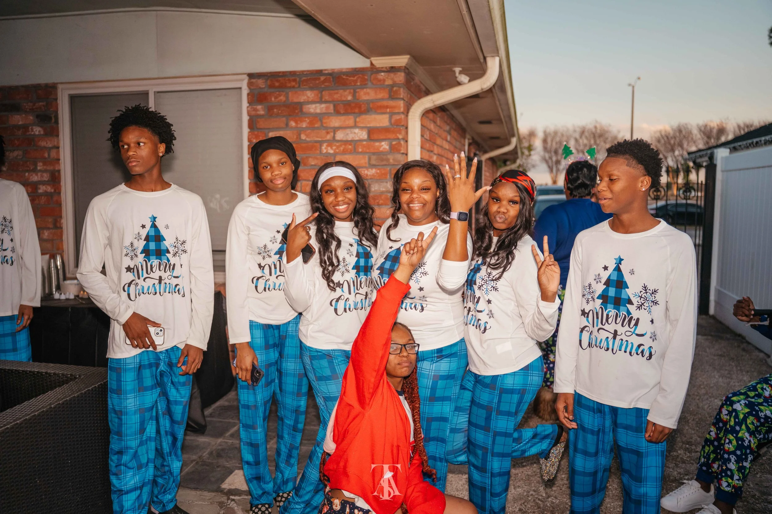 Group of children dressed in matching Christmas pajamas, some with holiday headbands, celebrating outside near a brick house during sunset.