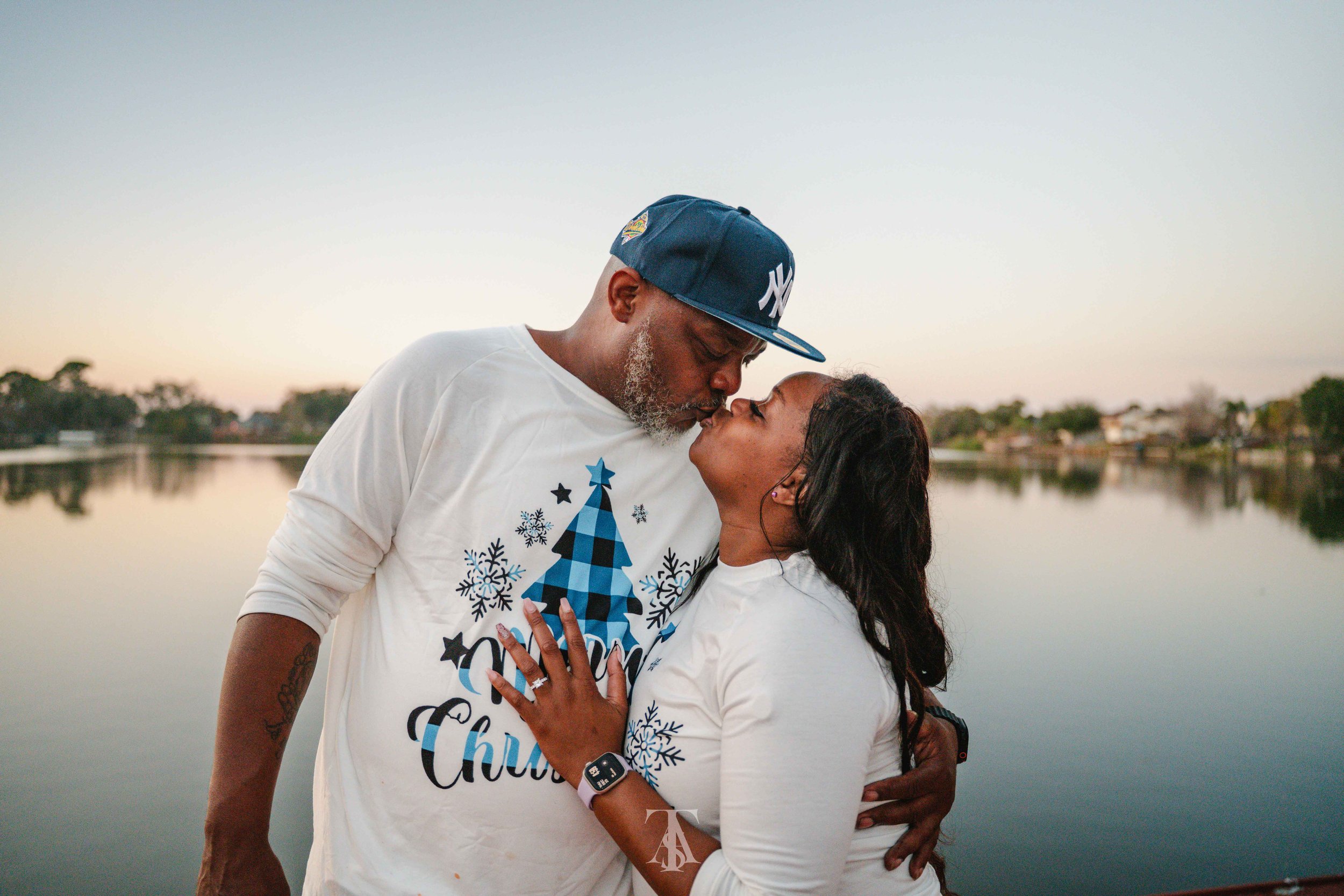 A couple is embracing near a body of water during sunset or sunrise, with their faces close together. They are wearing matching white holiday shirts with blue Christmas trees and snowflakes, and the woman has her hand on the man's chest. The man is w