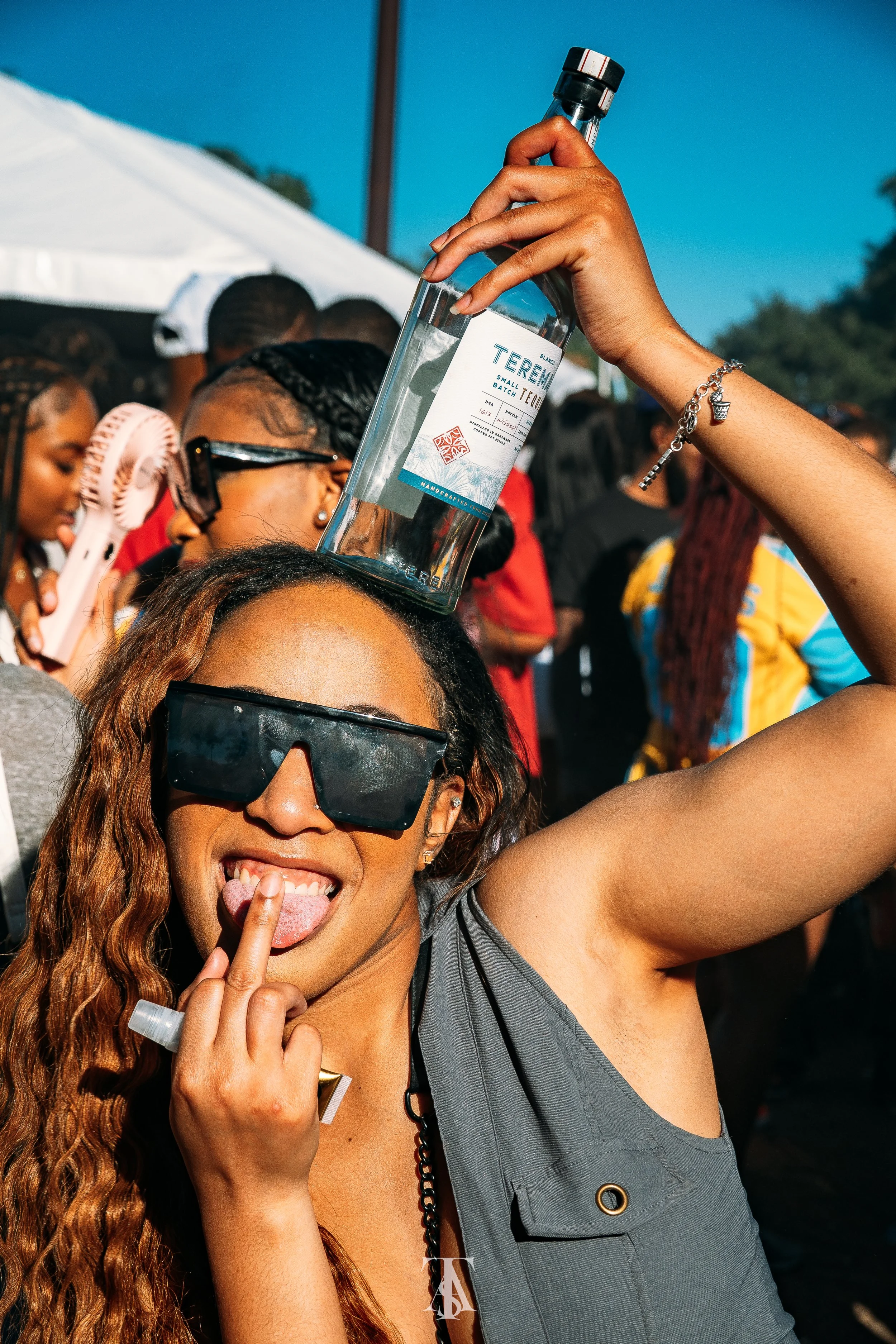 A woman with long curly hair and large sunglasses holding a bottle of vodka on her head at an outdoor event with a crowd of people in the background.