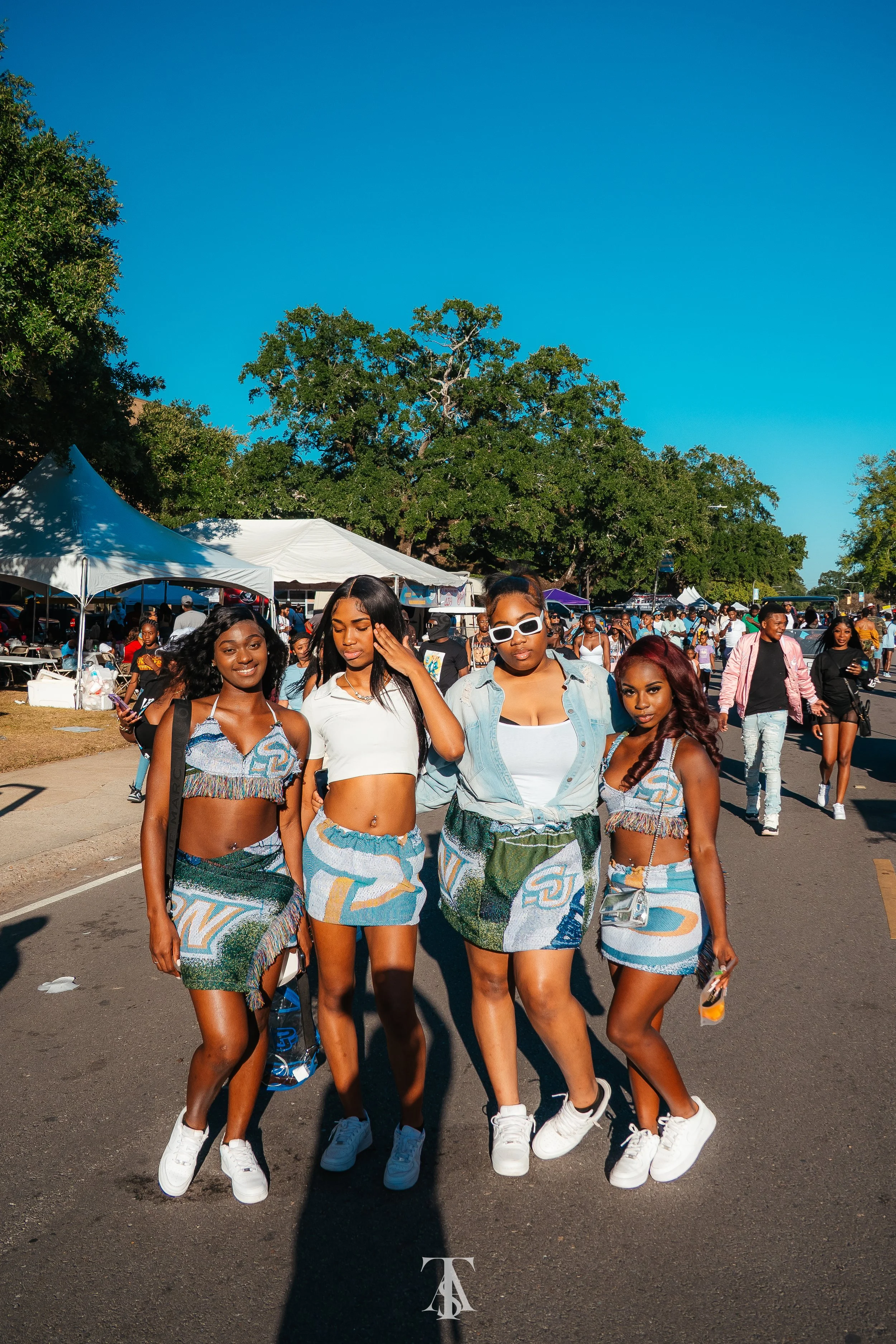 Four young women in colorful outfits walking together at an outdoor event on a sunny day with tents and other people in the background.