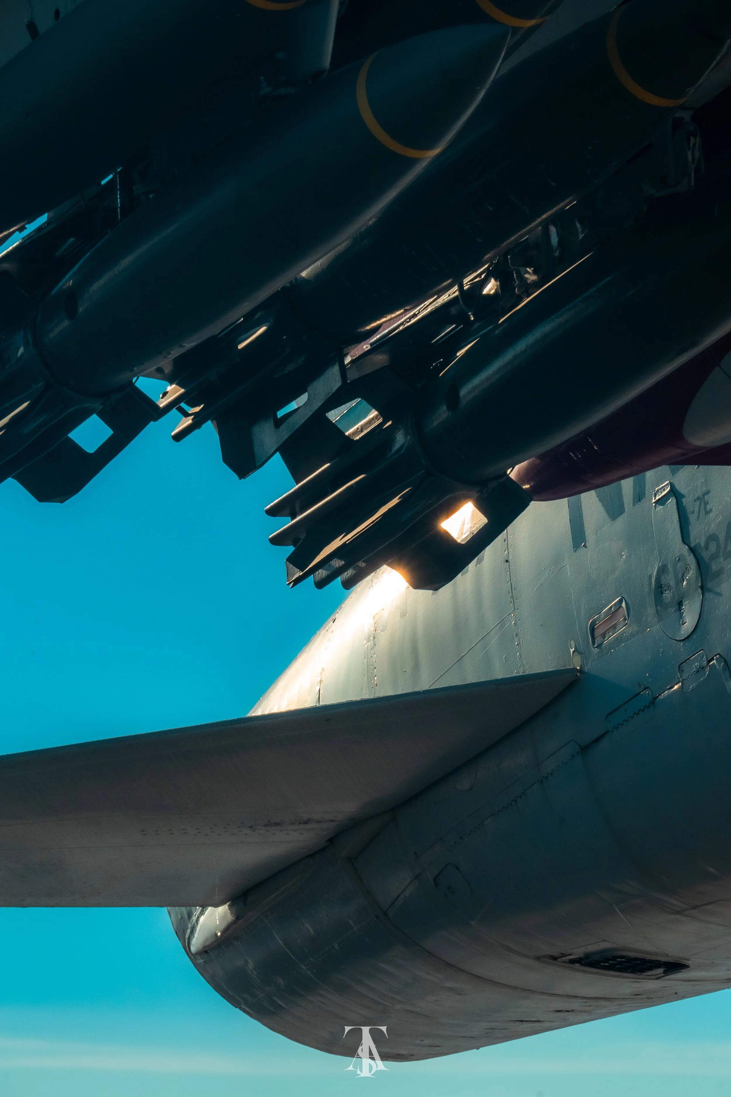 Close-up of a military aircraft's underbelly, showing exposed missile hardware against a blue sky.