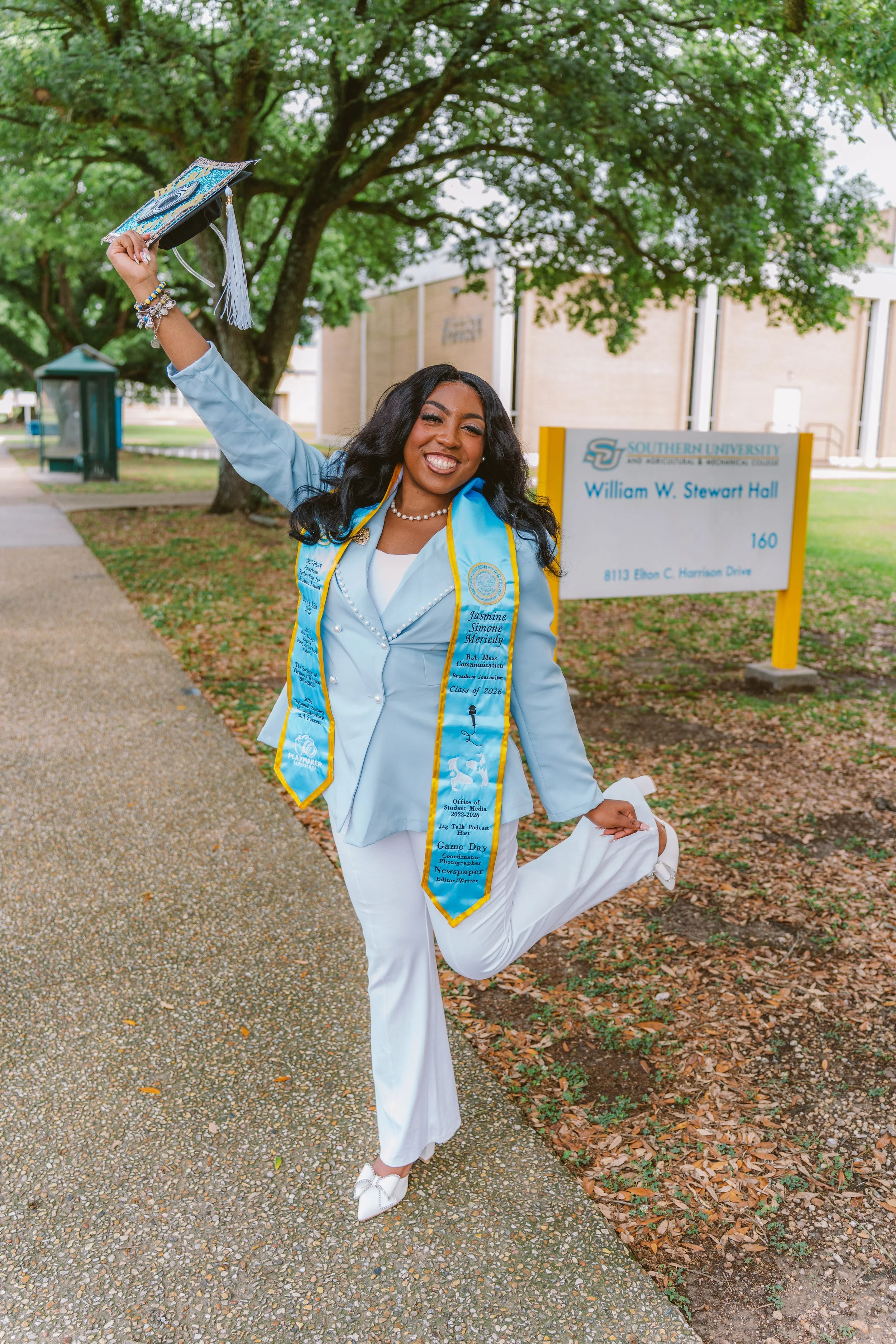 A young woman in graduation gown and sash, smiling and holding her cap in the air, standing outside on a pathway with trees, near a campus sign that reads "William W. Stewart Hall," Southern University.