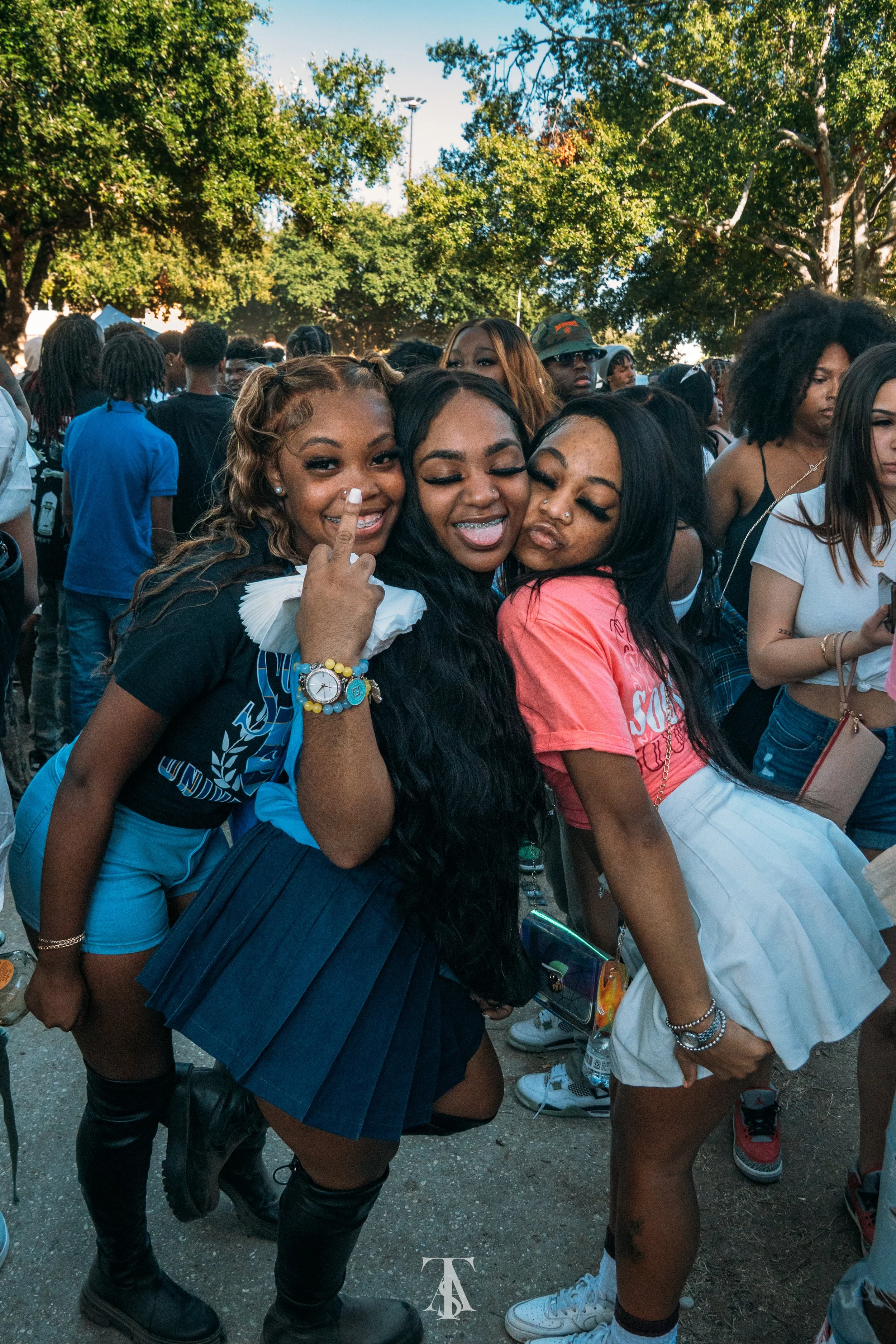 Three young women smiling and posing for a photo at an outdoor crowd event, with trees in the background.