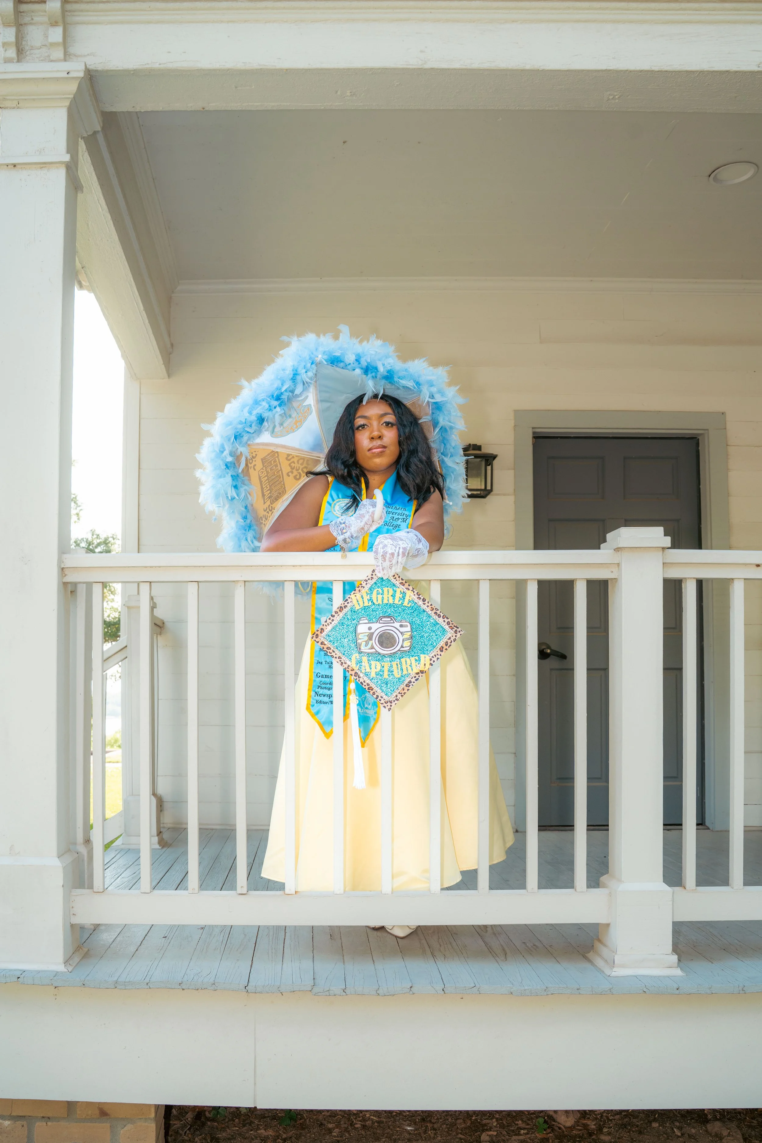 A woman standing on a porch, wearing a yellow dress, a blue sash, and white gloves, with a decorative umbrella hat and holding a ribbon that says 'Graduate' and features a camera graphic.