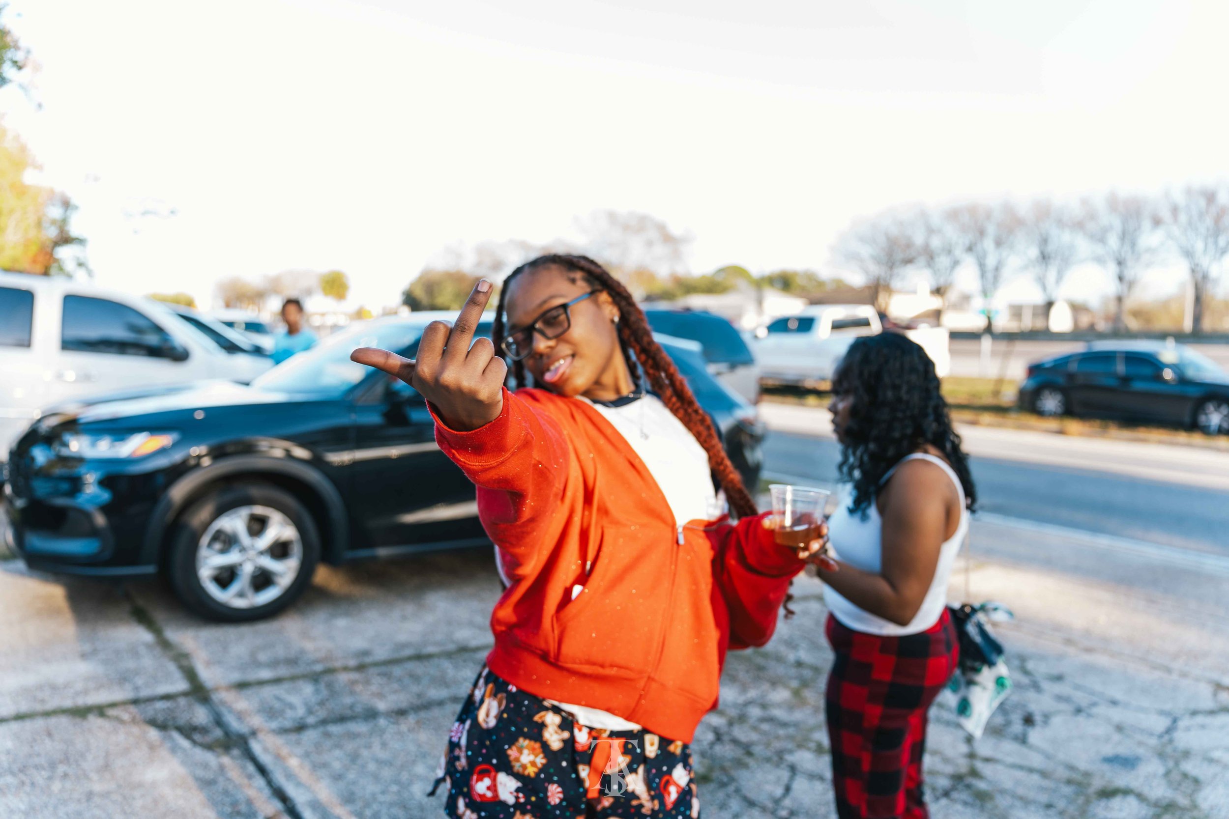 A woman with glasses making a hand gesture in front of her face, holding a drink, and smiling outdoors with other people and cars in the background.