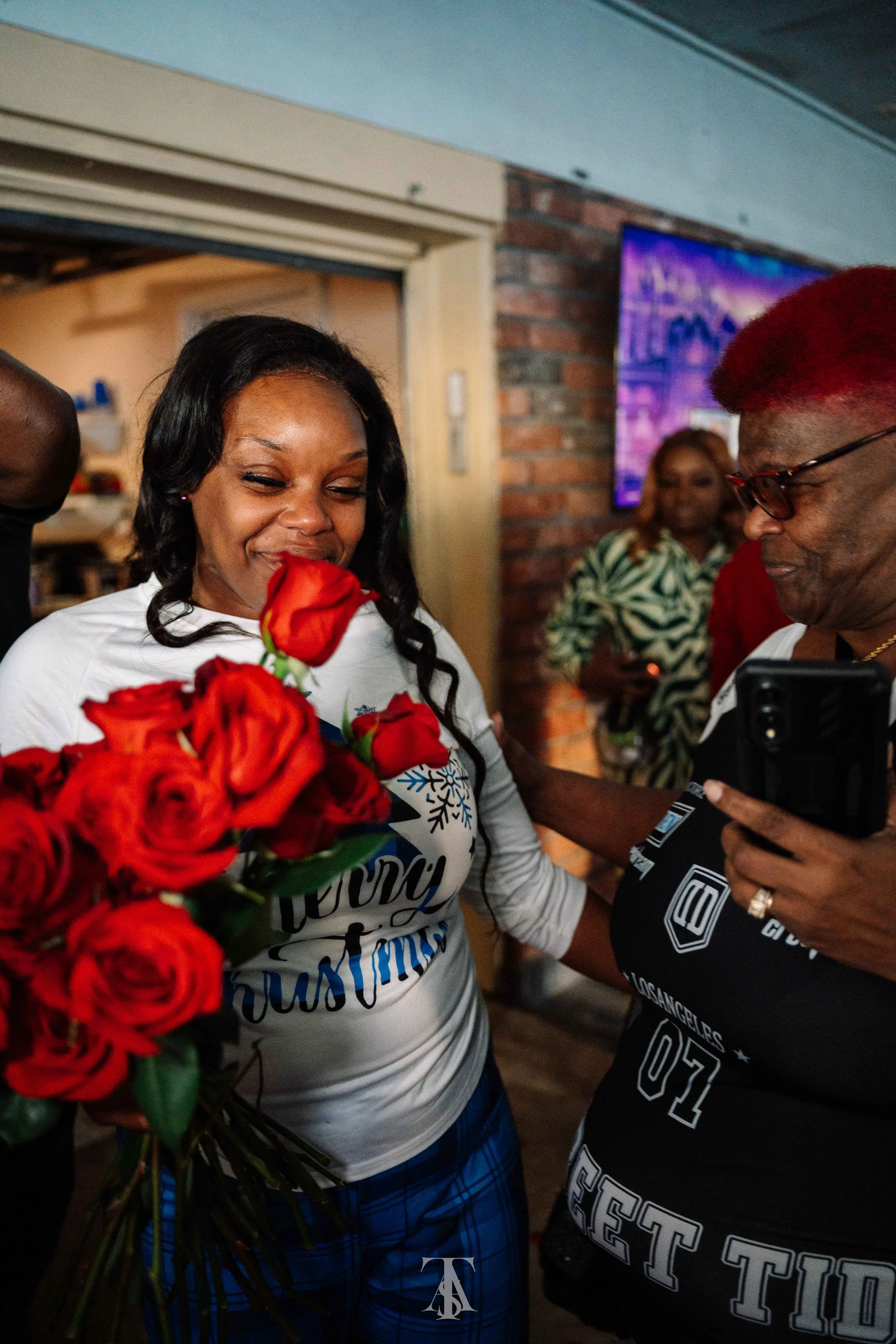 A woman with long black hair holding a bouquet of red roses, smiling with eyes closed, wearing a white shirt with blue writing, blue plaid shorts, and surrounded by people in a room with brick walls and a TV screen in the background.