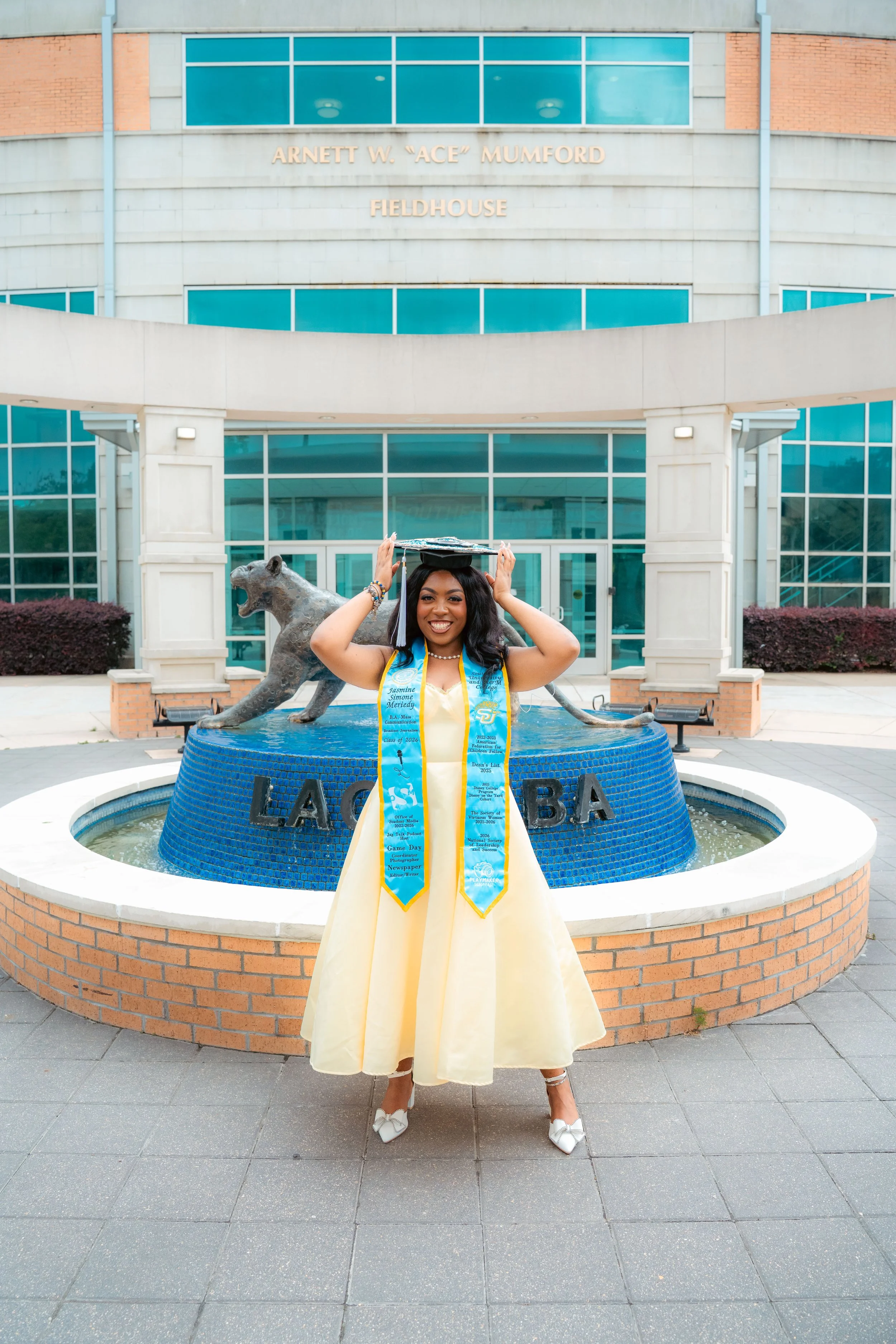 A young woman in a yellow dress with white shoes and graduation regalia holding a graduation cap above her head, standing in front of a blue and brick building, with a statue of a panther and a fountain with the words 'LACLEDA BANK' written around it