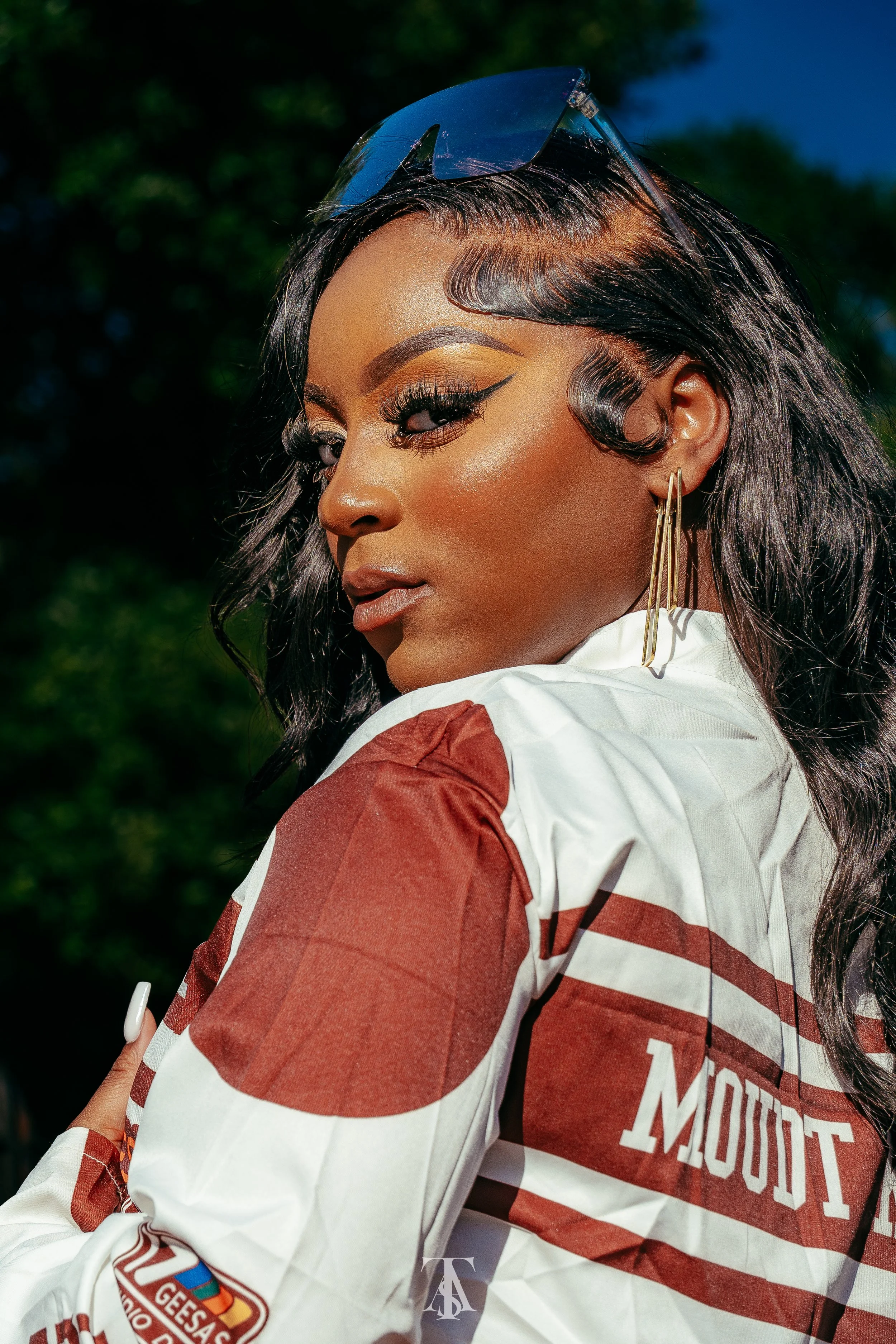 A woman with dark wavy hair, sunglasses on her head, makeup, and gold earrings, wearing a racing jacket with patches, posing outdoors.